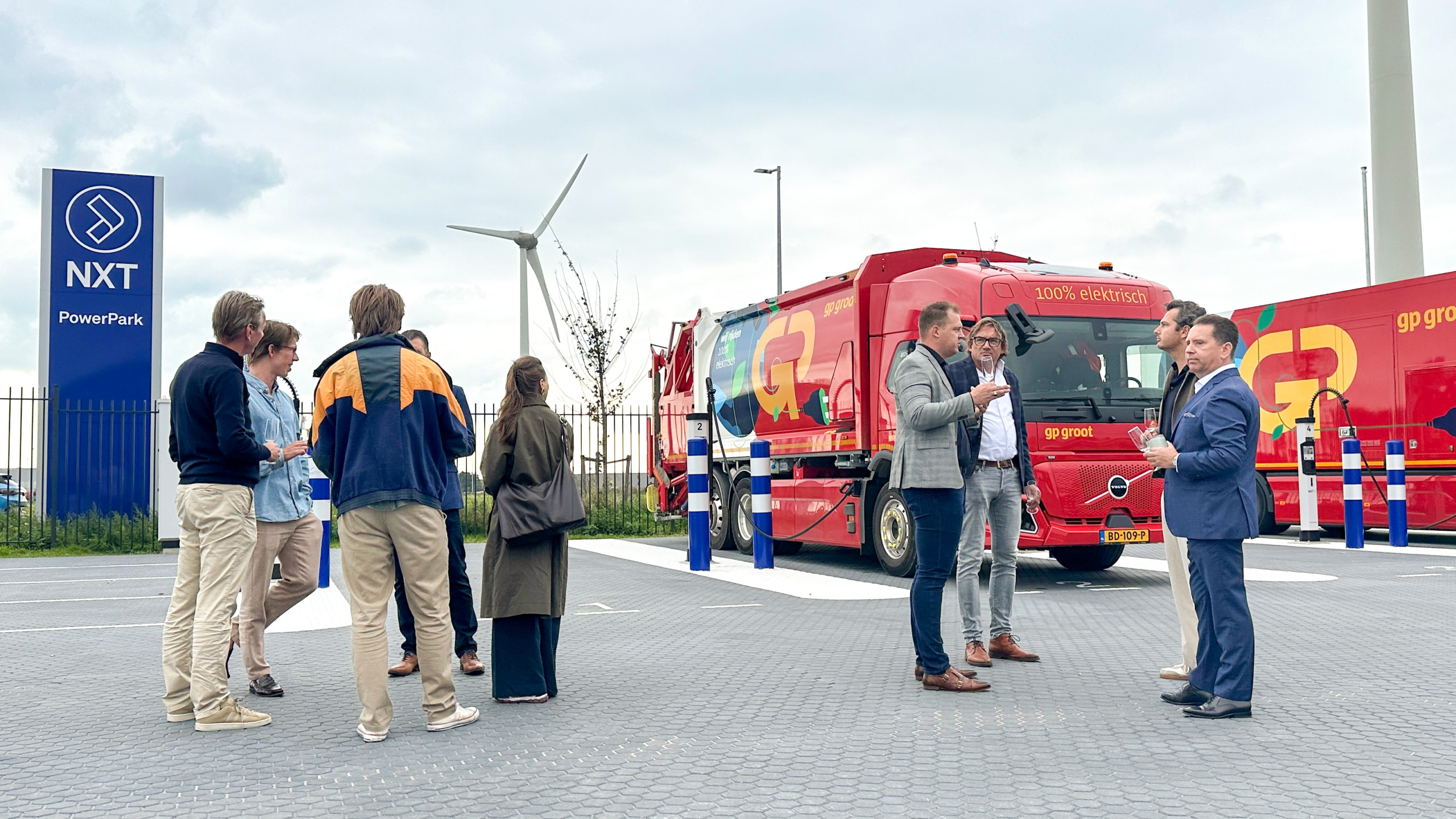 Groep mensen praat bij een laadpunt naast een rode vrachtwagen met tekst 100% elektrisch en een windturbine op de achtergrond