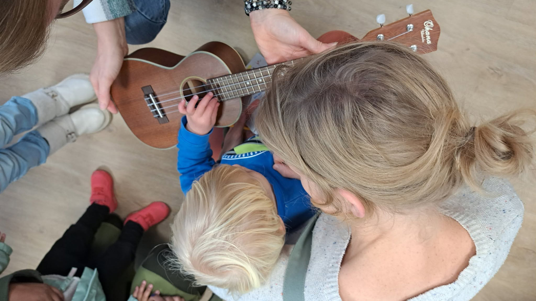 Van bovenaf: volwassene helpt kind een ukelele vasthouden en spelen, met andere kinderen eromheen