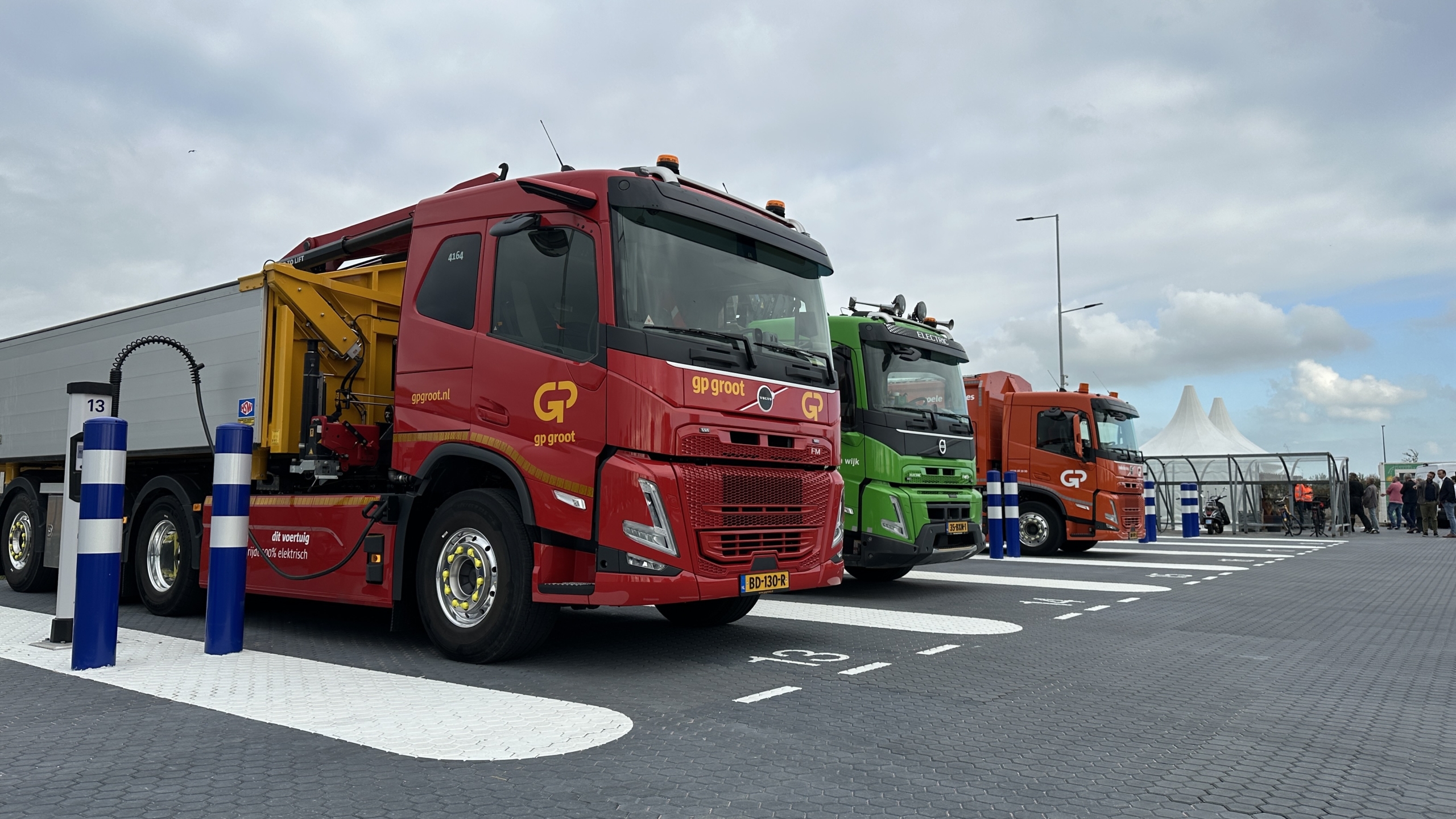 Drie vrachtwagens (rood, groen en oranje) geparkeerd op een terrein met blauw-witte paaltjes onder bewolkte lucht