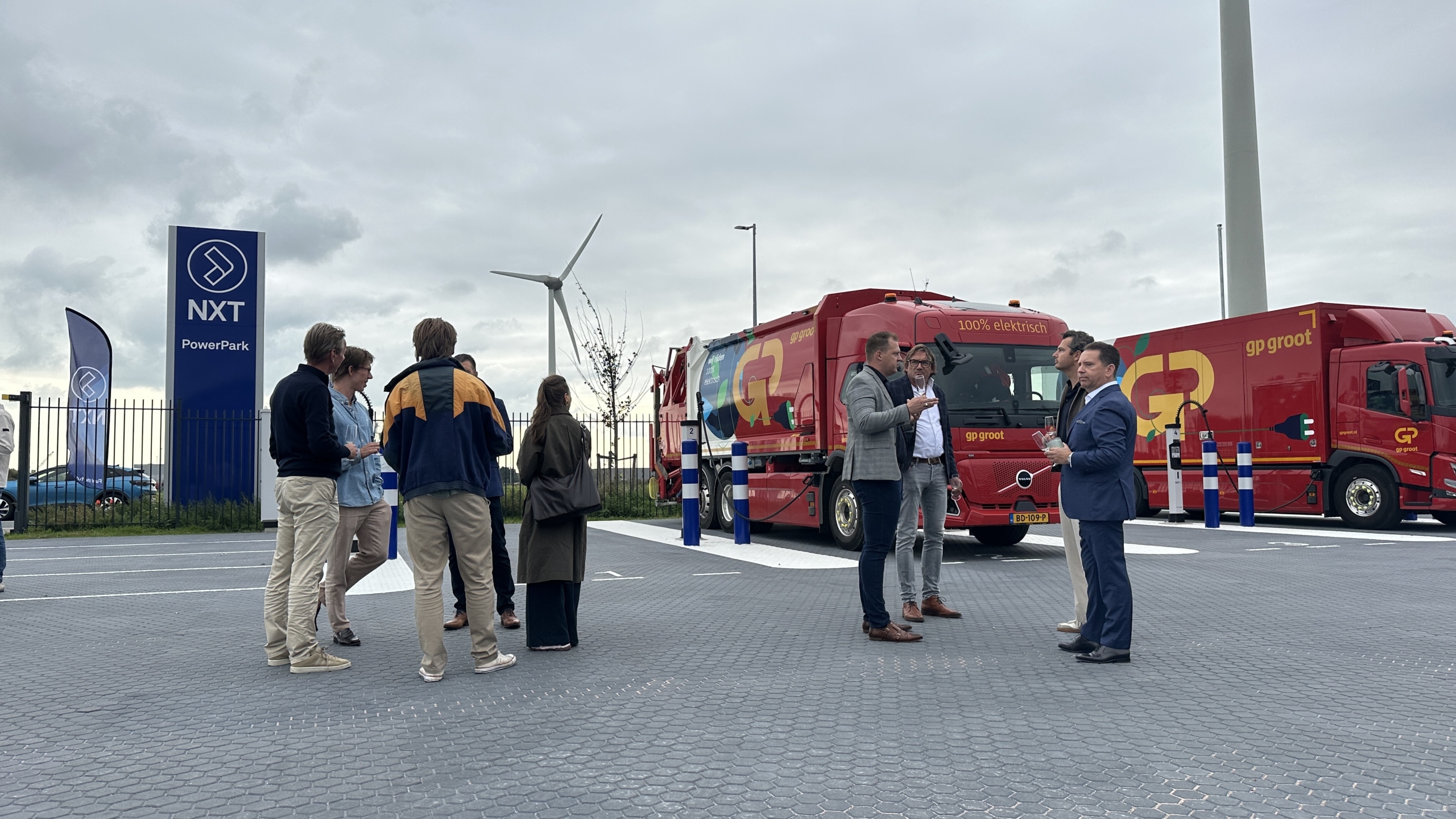 Groep mensen praat bij NXT PowerPark naast rode elektrische vrachtwagens van gp groot; windturbine op de achtergrond