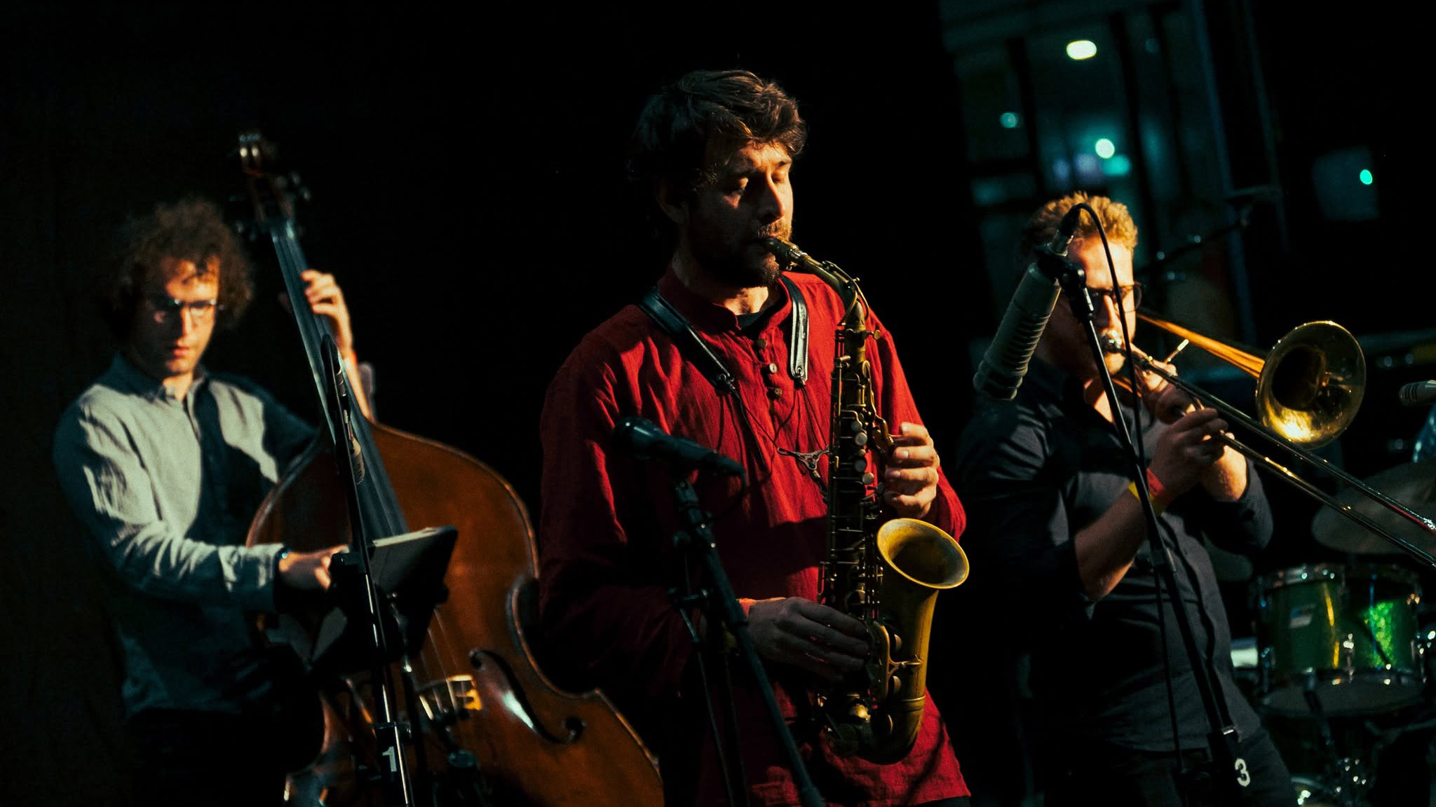 Jazztrio op een donker podium: contrabas links, saxofonist in rood midden en trombonist rechts onder spotlicht