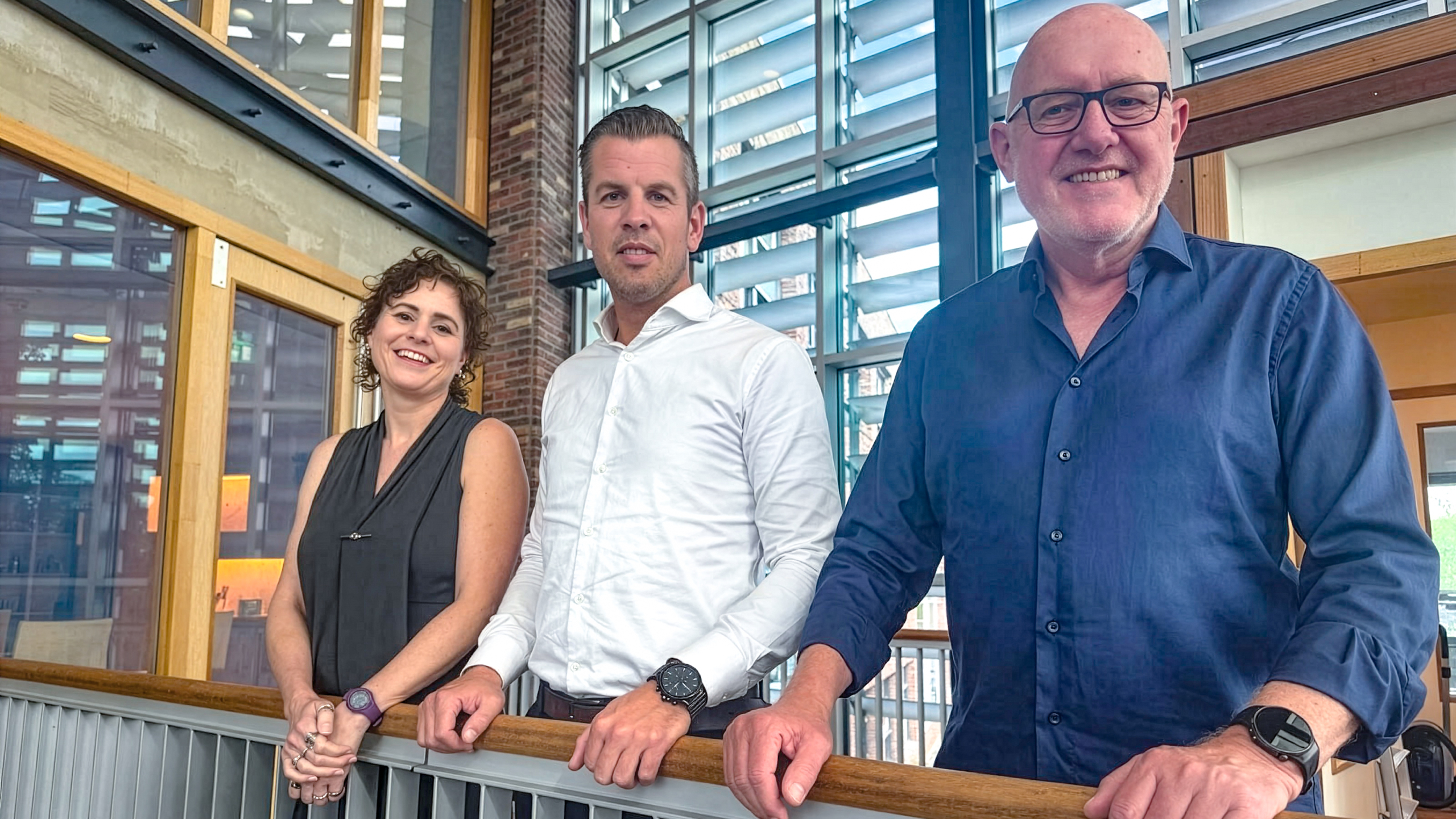 Drie collega’s leunen op een balustrade in een modern kantoor met grote ramen en houten details