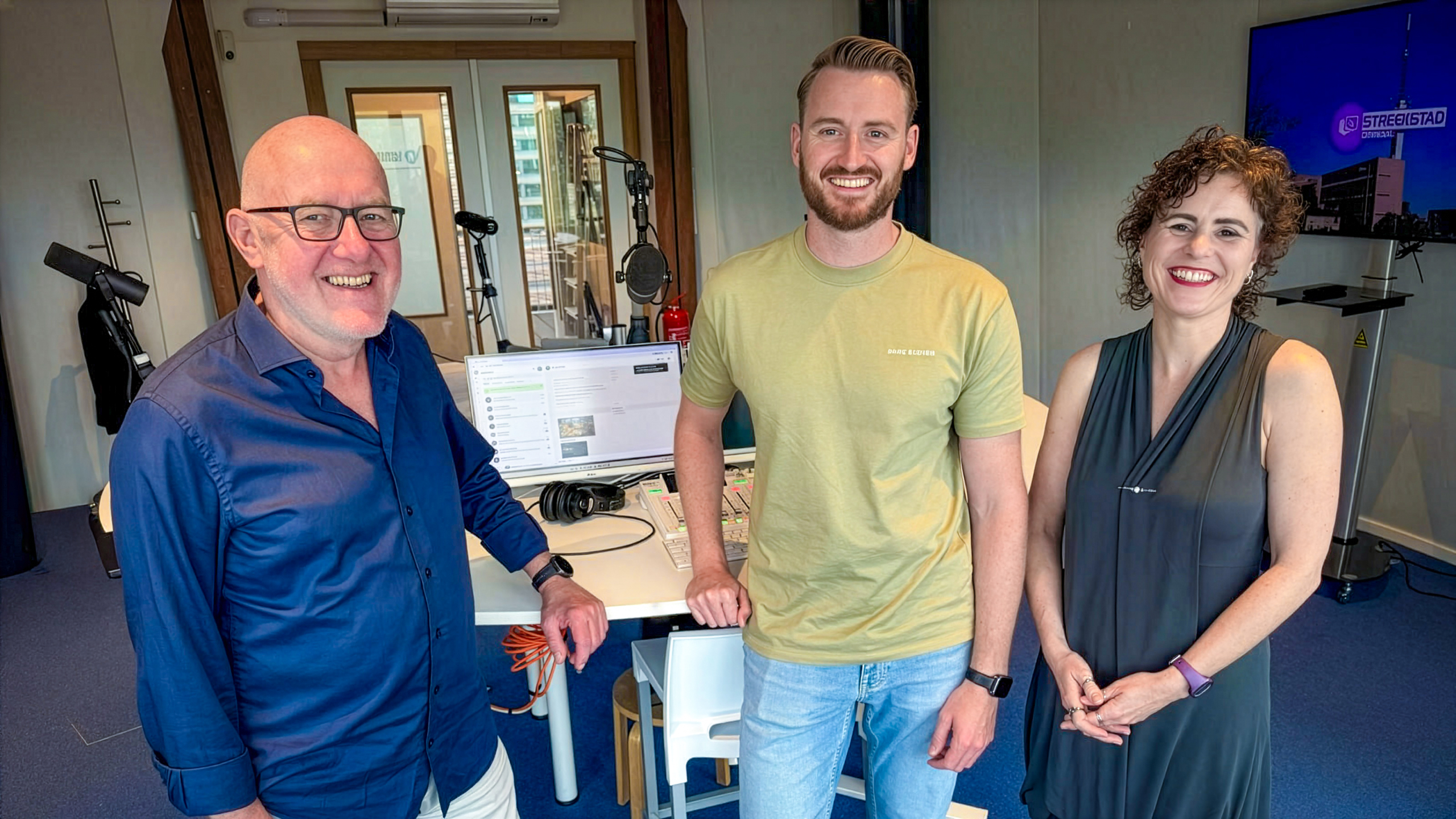 Drie lachende mensen in een radiostudio bij een tafel met microfoons, mengpaneel en computer