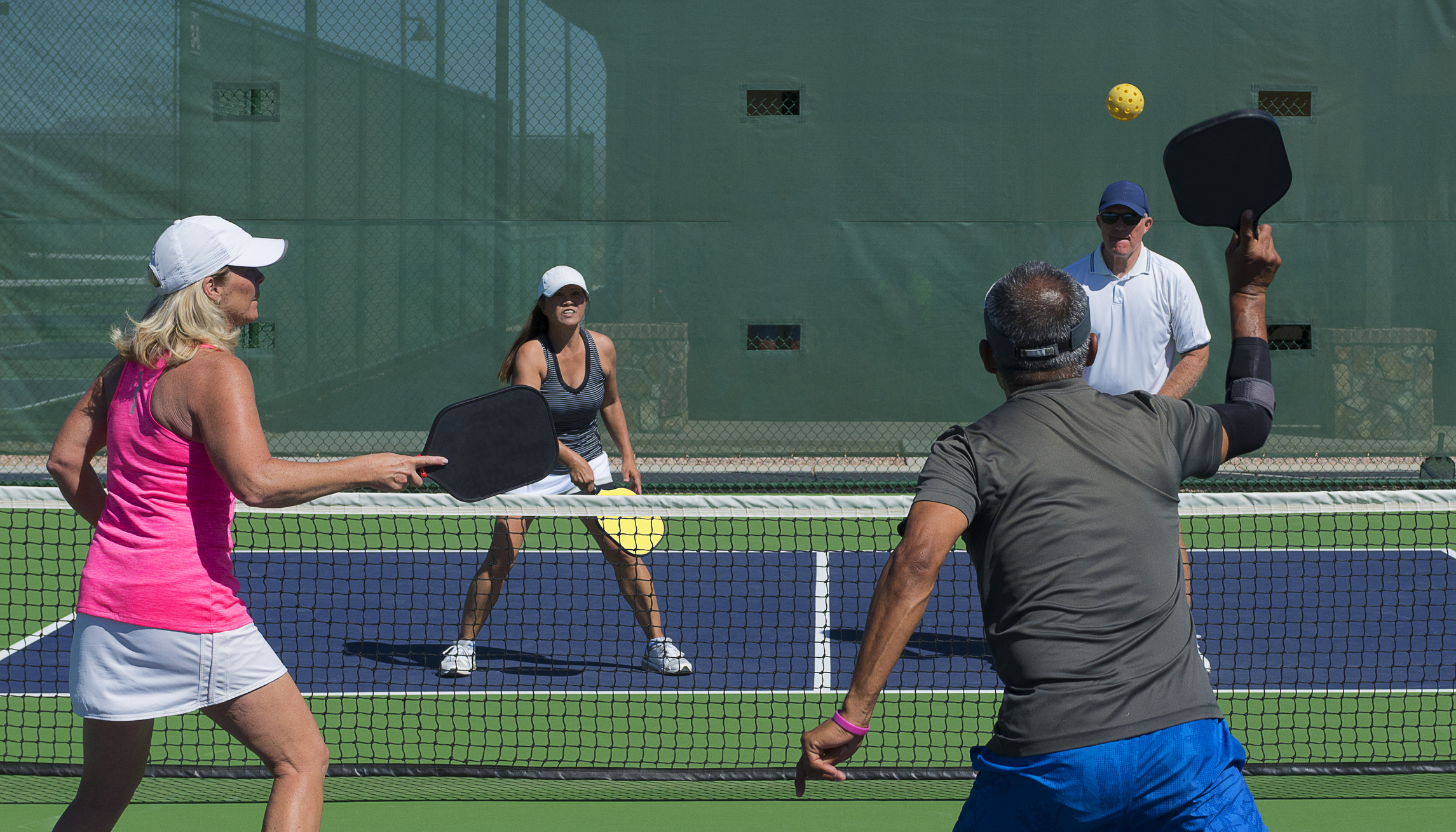 Vier spelers spelen pickleball op een buitenbaan, met paddles omhoog terwijl een gele bal over het net vliegt