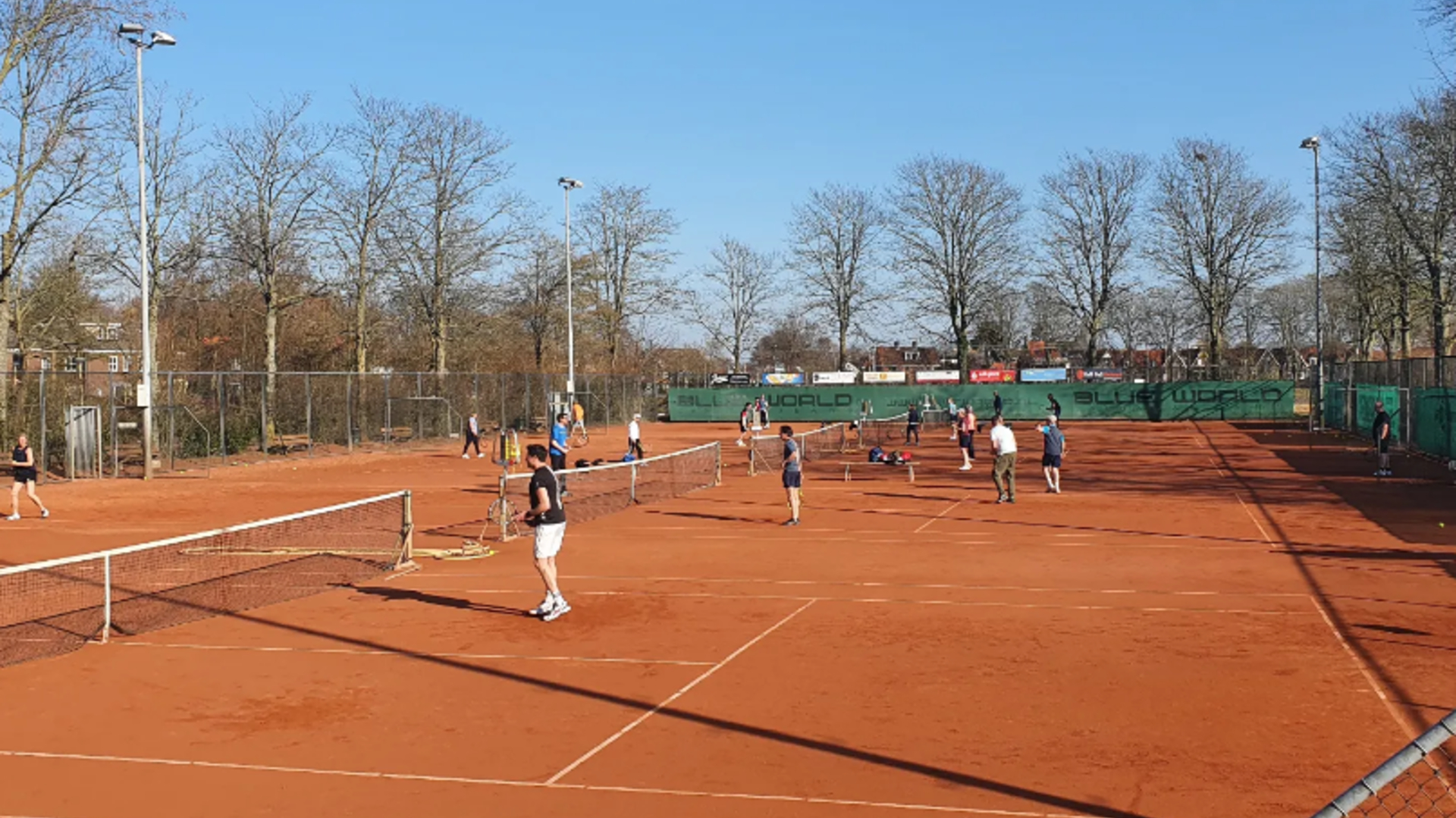 Mensen spelen tennis op meerdere gravelbanen buiten, met netten, hekwerk en kale bomen onder een blauwe lucht