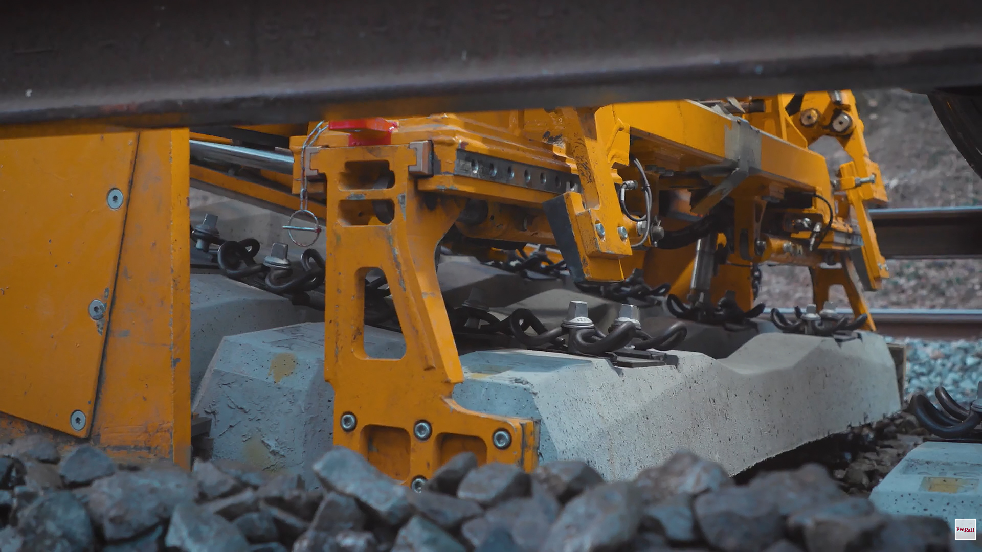 Gele spoorbouwmachine bevestigt een betonnen dwarsligger met klemmen en bouten tussen het ballastgrind