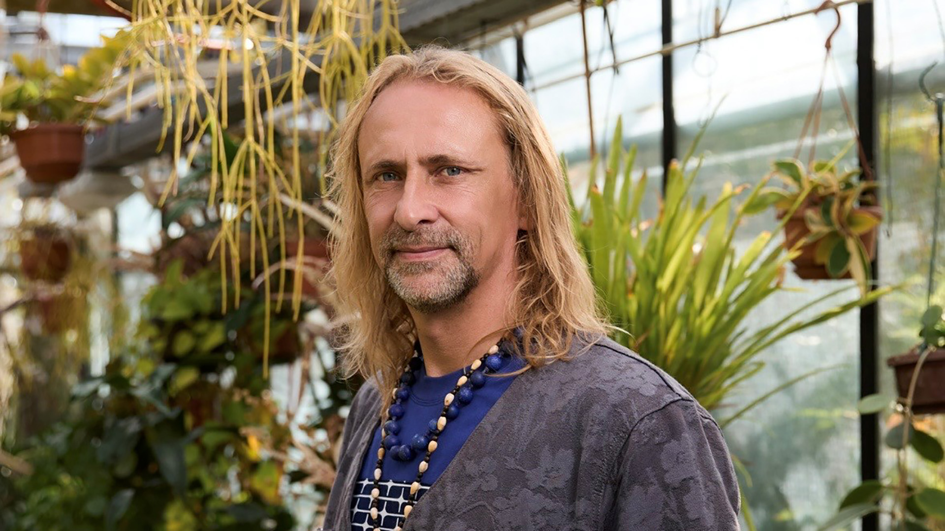 Portret van een man met lang blond haar en baard, staand in een kas tussen hangende planten