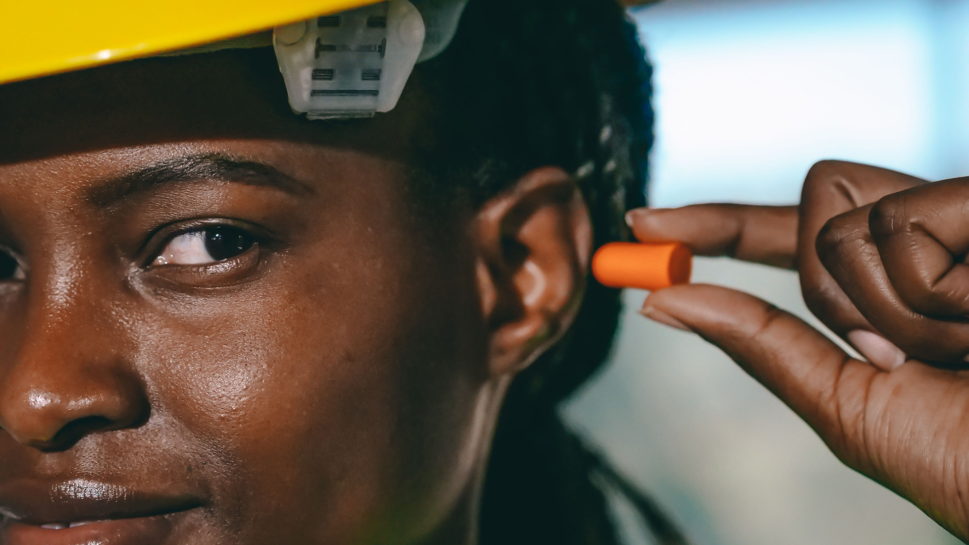 Close-up van een bouwvakker met gele helm die een oranje oordopje in het oor doet