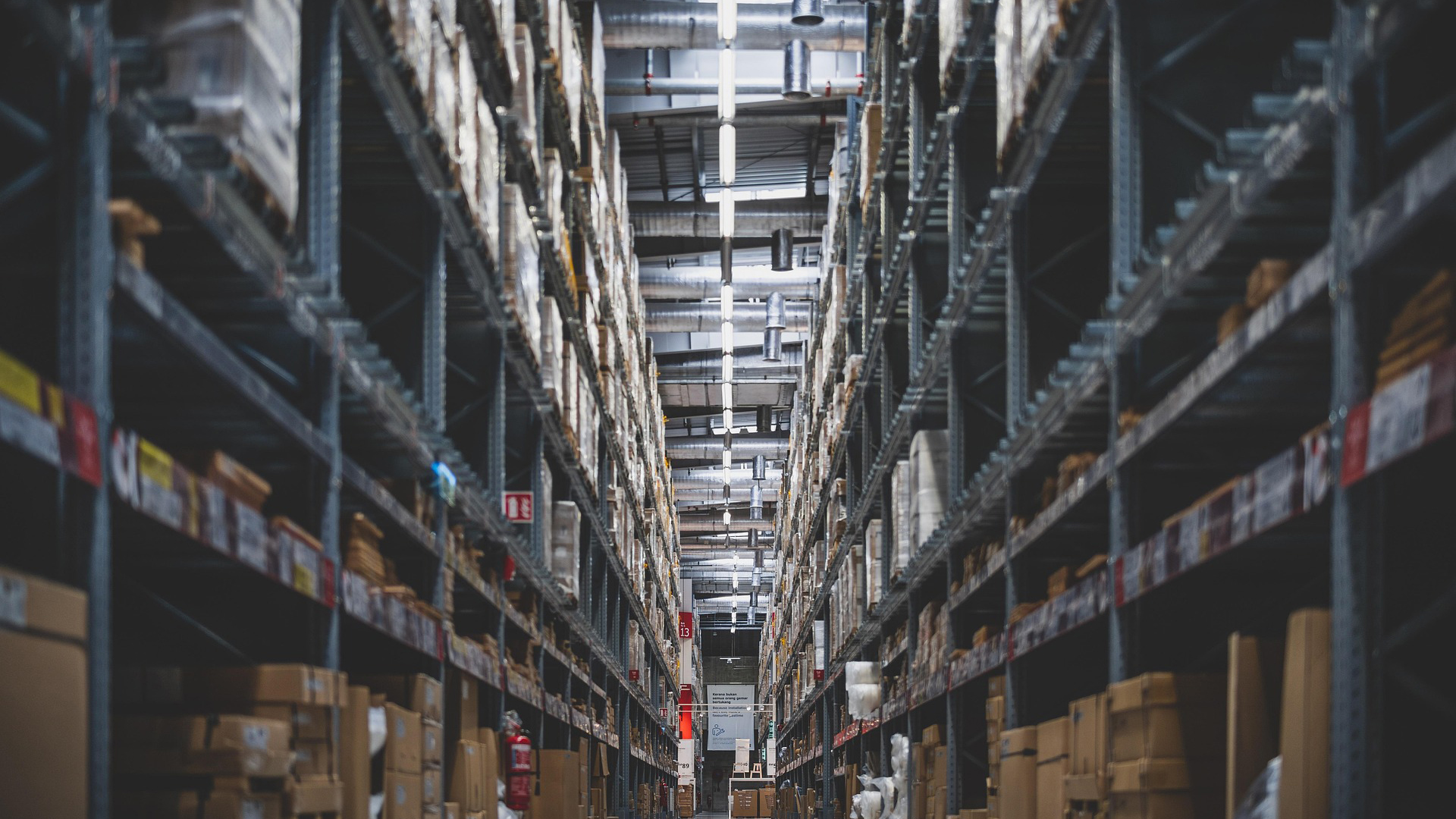 Lange gang in een magazijn met hoge stellingen vol dozen en pallets, met TL-verlichting en ventilatiebuizen aan het plafond