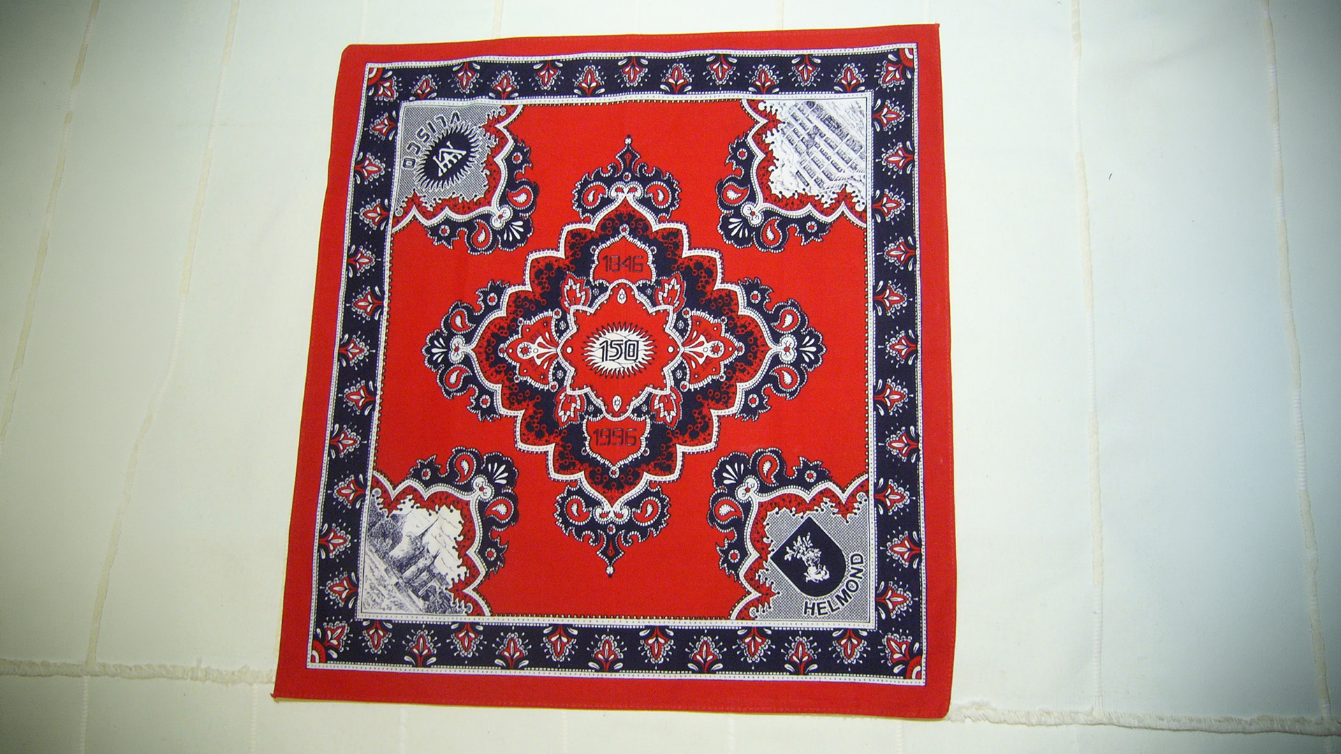Rode bandana met zwart-wit paisleypatroon en centrale medaillon, met tekst “HELMOND” en jaartallen 1946 en 1996
