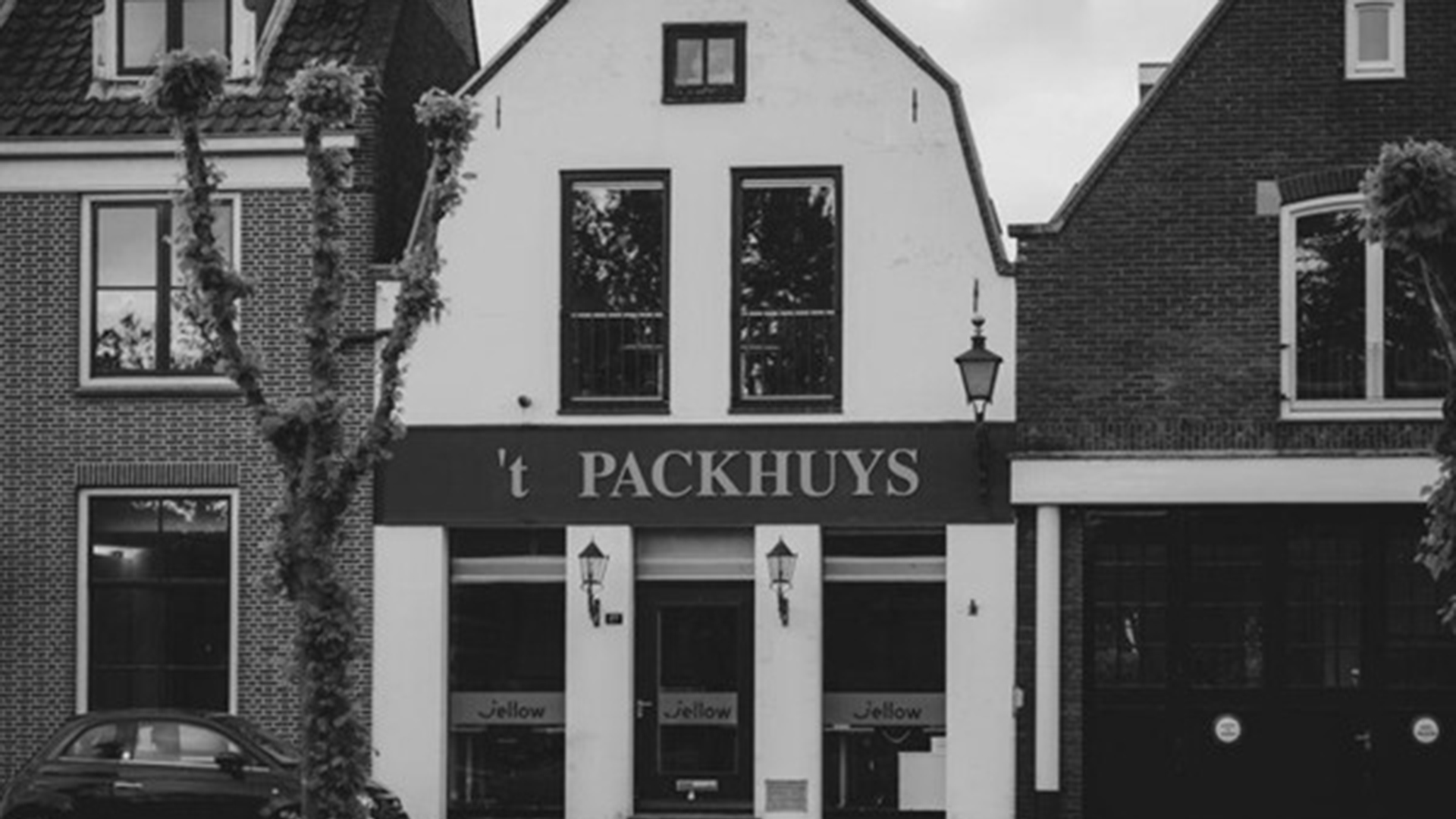 Zwart-witfoto van gevel met opschrift ’t PACKHUYS, ramen en lantaarns, met een geparkeerde auto ervoor