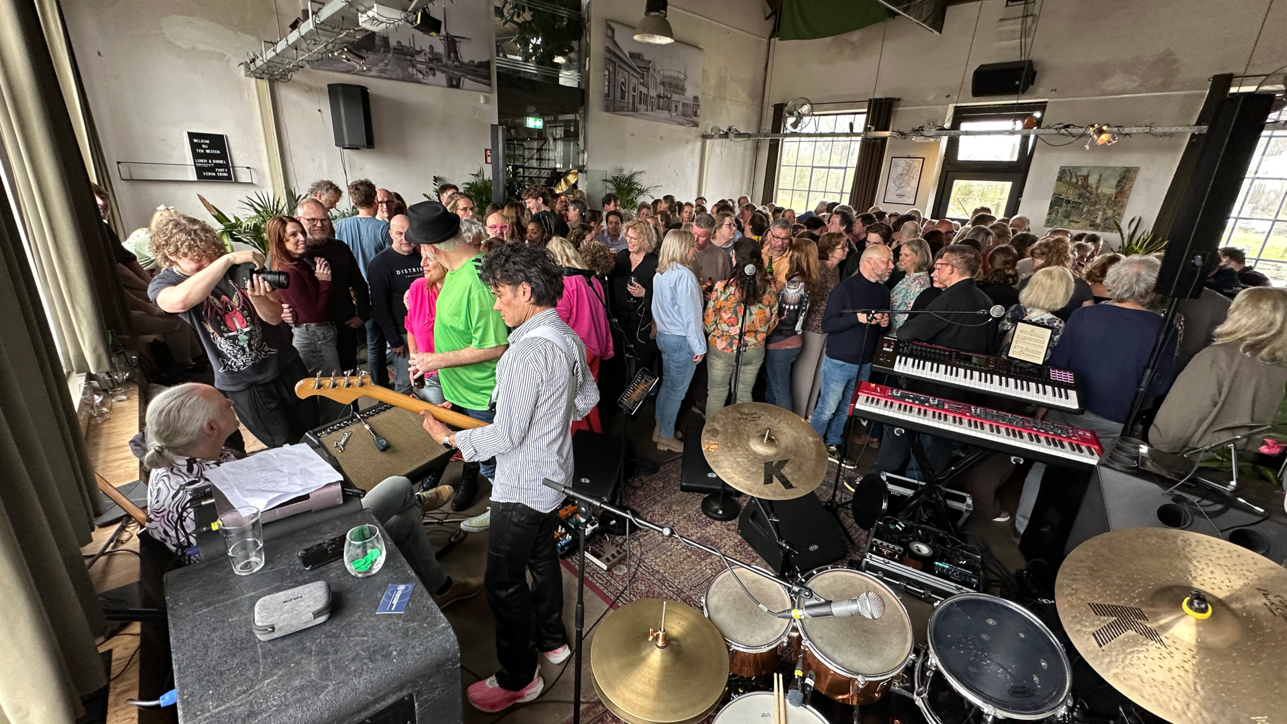 Drukke zaal met publiek rond het podium met drumstel, keyboards en bassist tijdens een liveoptreden