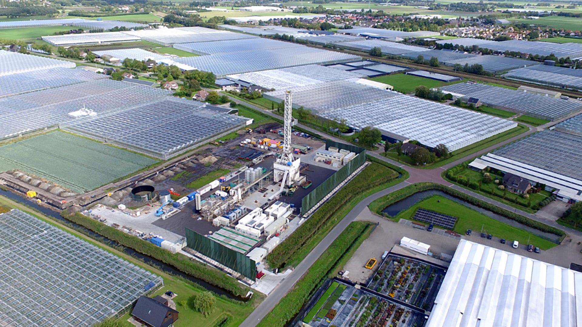 Luchtfoto van groot kassencomplex met industriële boortoren en bouwplaats, omringd door wegen, sloten en akkers