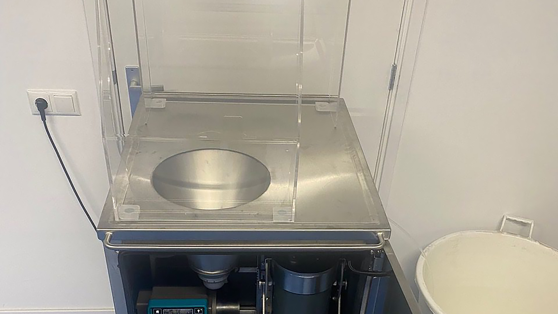 Industriële machine met transparante kap en ronde metalen kom op een werkblad, met open onderkast en opvangbak ernaast