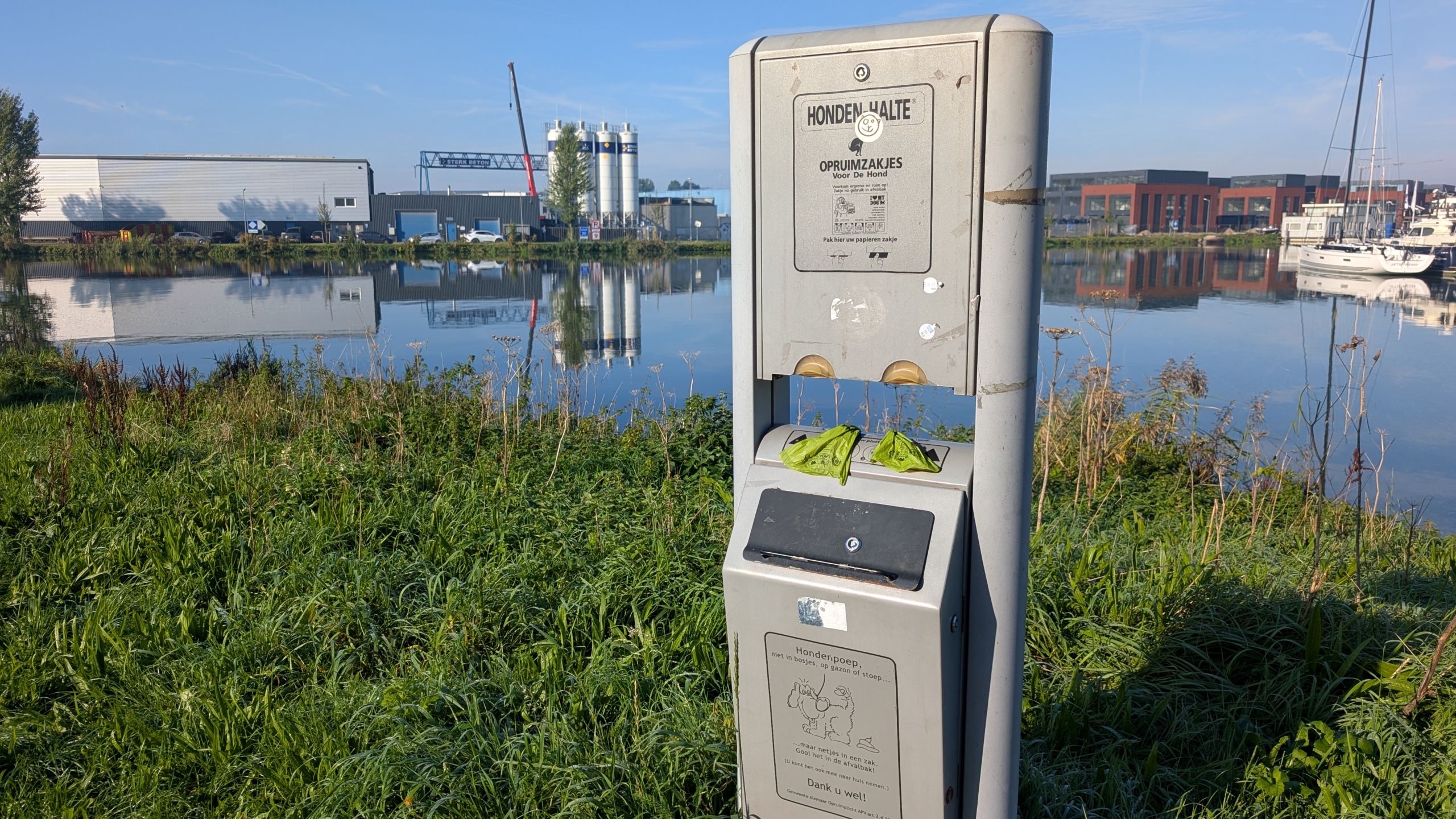 Hondenpoepzuil met opruimzakjes bij een waterkant, met industriegebouwen en zeilboten op de achtergrond