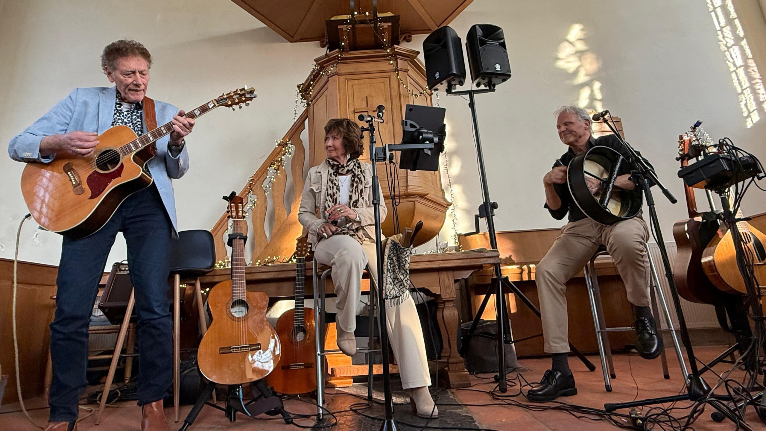 Muzikanten op klein podium: man speelt akoestische gitaar, vrouw zit tussen microfoons, man speelt handdrum in kerkruimte