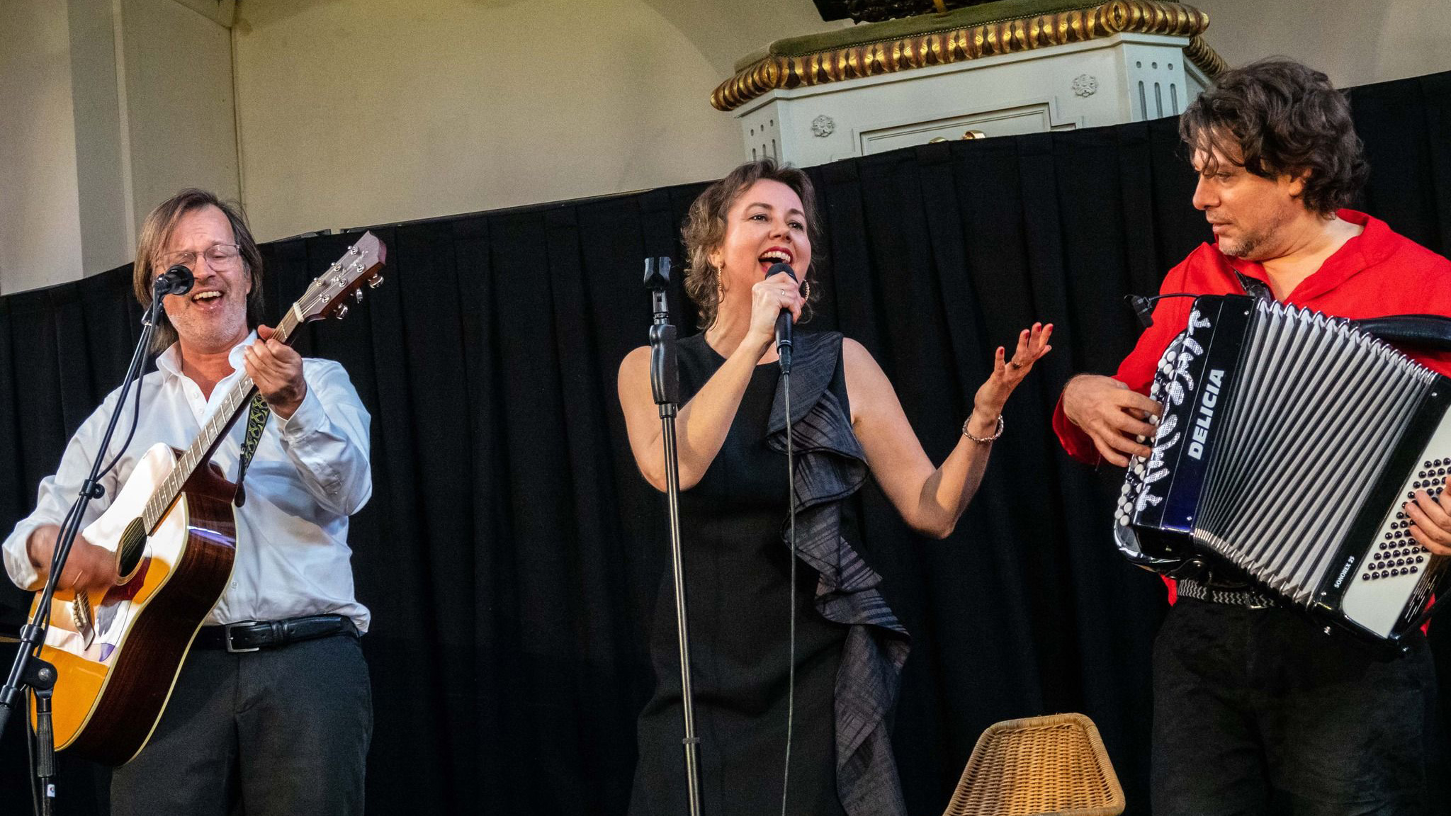 Drie muzikanten op een podium: zangeres met microfoon, gitarist links en accordeonist rechts