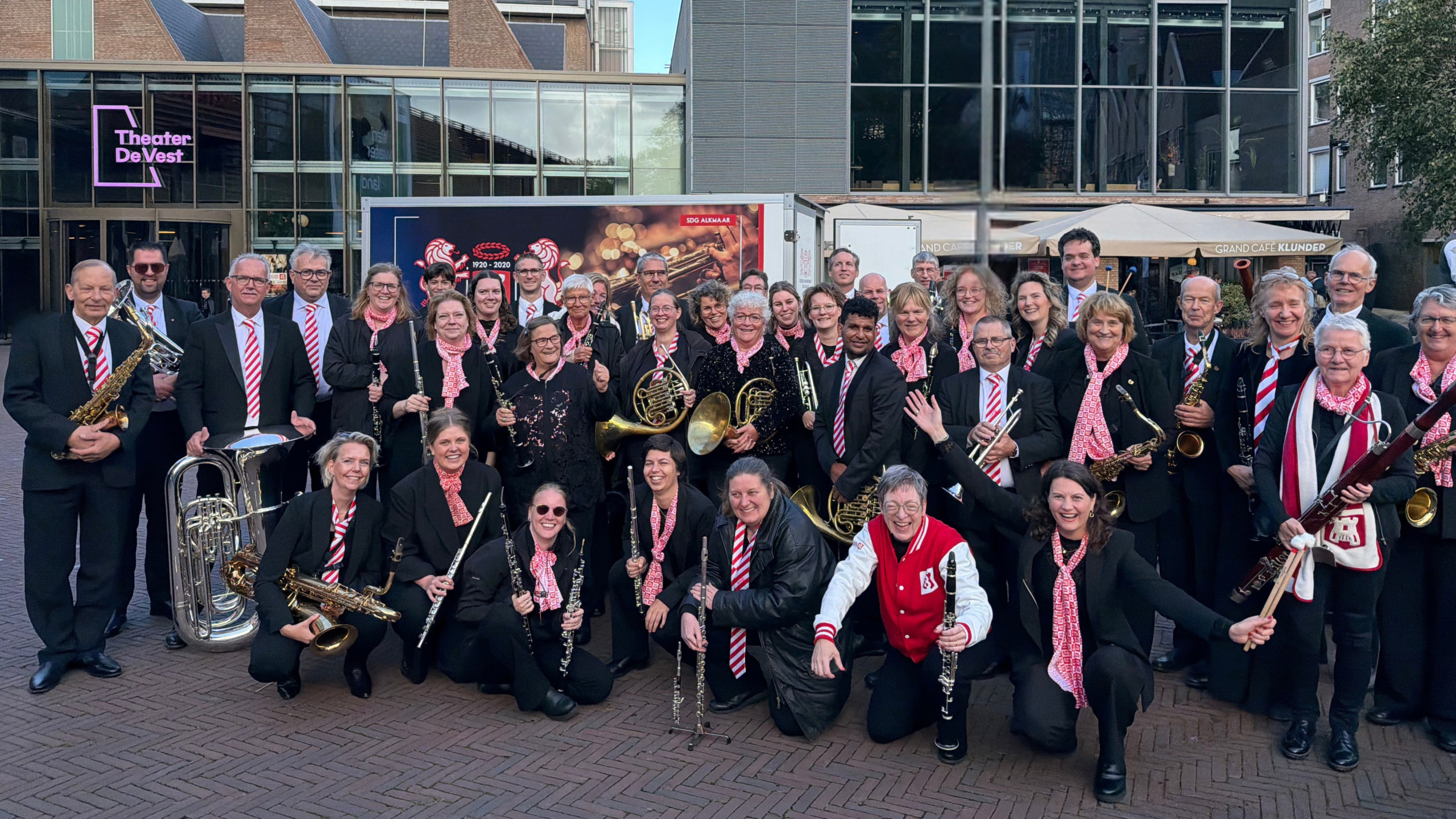 Groepsfoto van een orkest in zwarte kleding met rood-witte accenten en blaasinstrumenten voor Theater De Vest