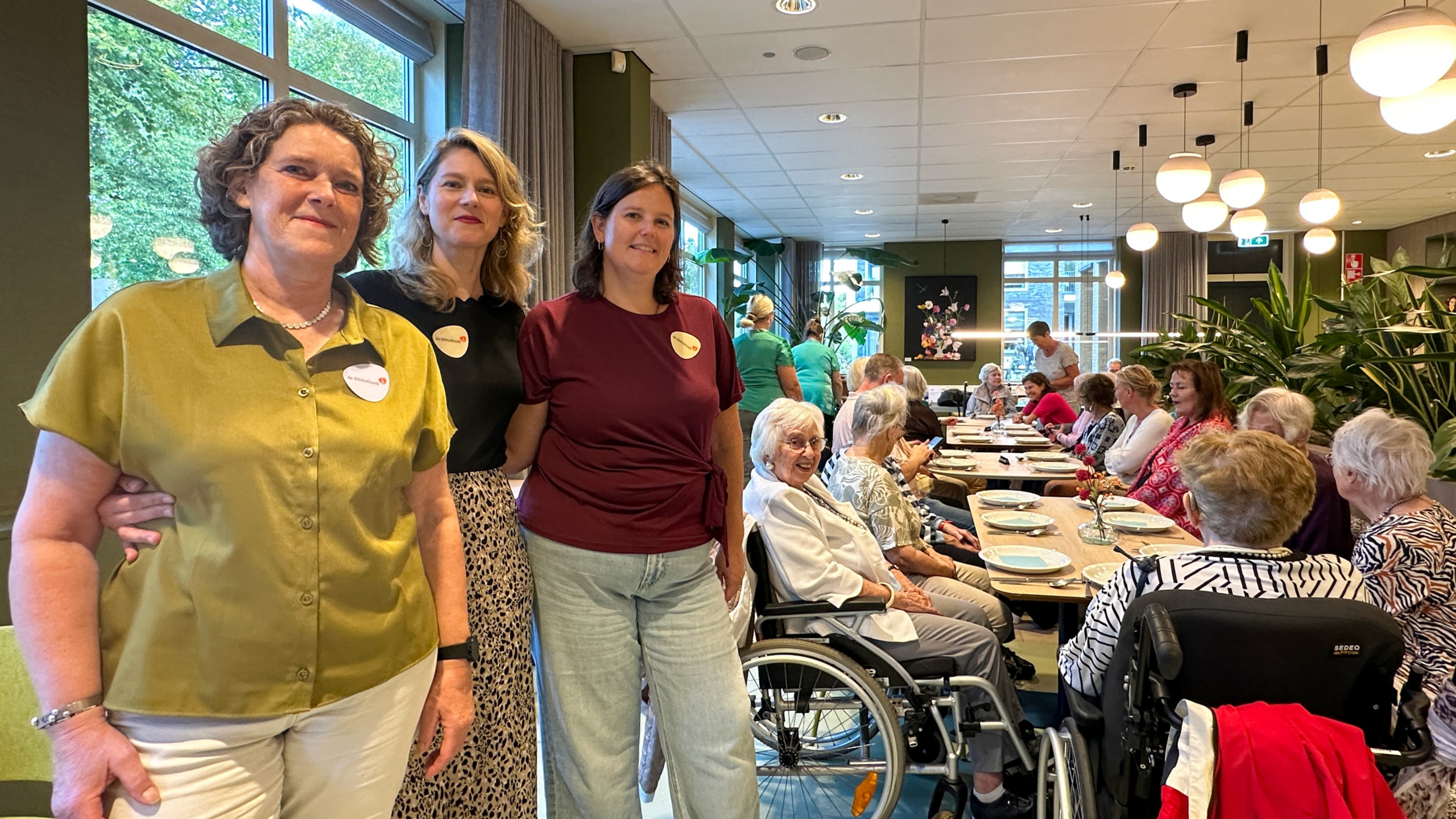 Drie vrouwen met naambadges poseren in een restaurantzaal waar ouderen aan lange tafels samen eten