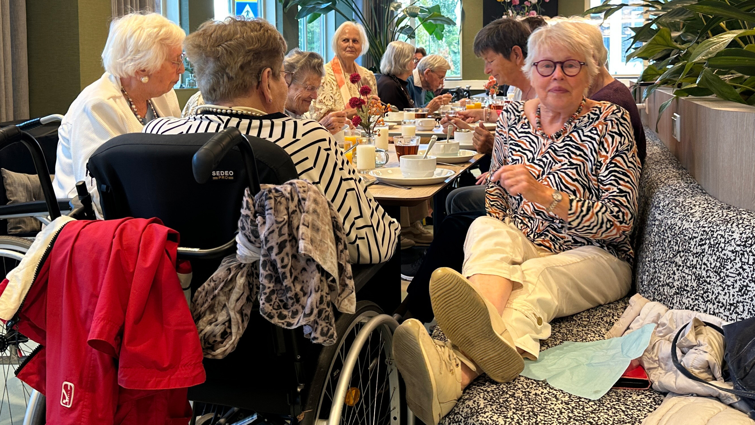 Groep oudere vrouwen aan een lange tafel in een restaurant, etend en pratend, met rolstoelen op de voorgrond