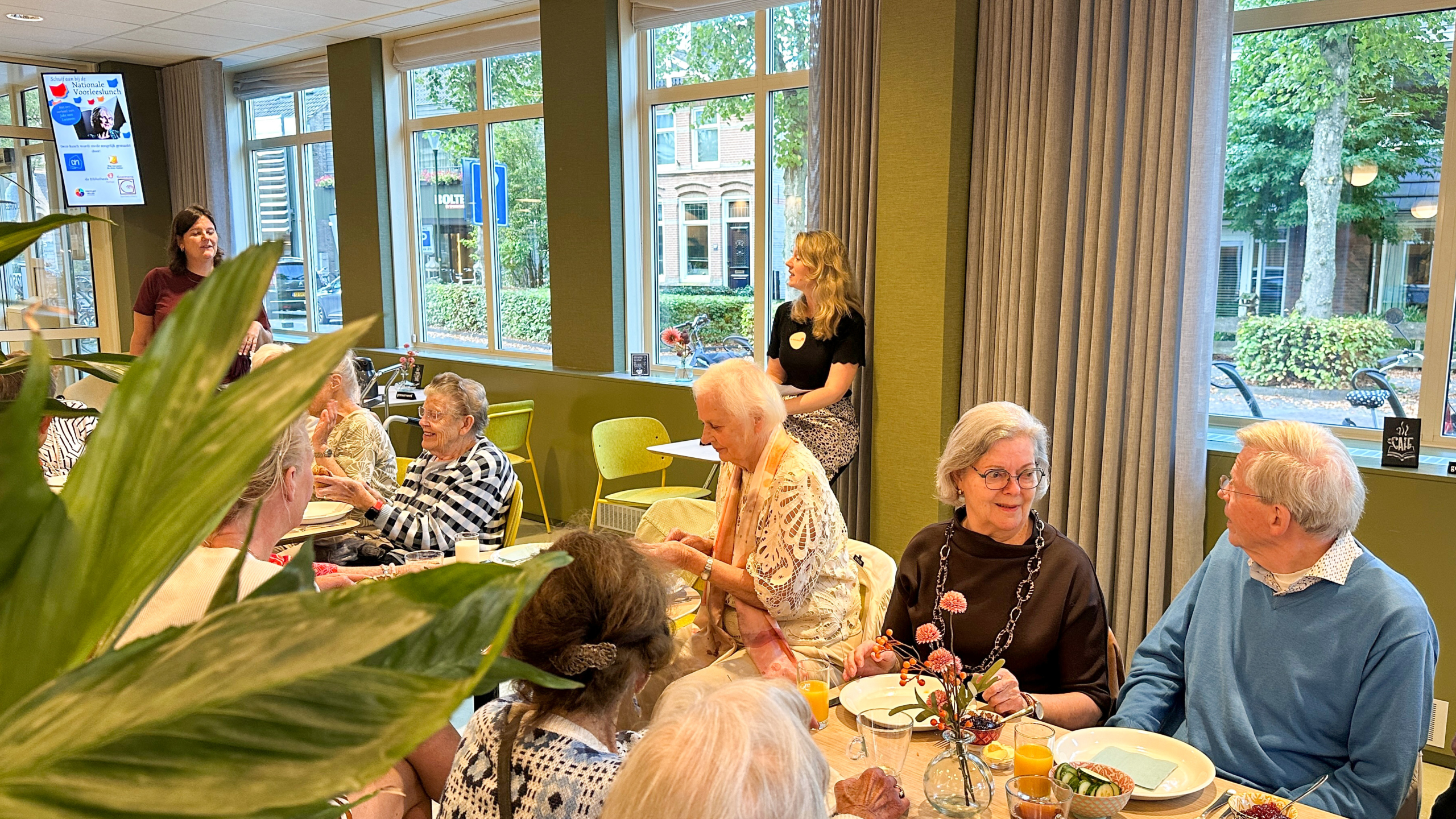 Groep ouderen aan tafel in een buurthuis, etend en pratend bij grote ramen; op de achtergrond twee vrouwen en een scherm