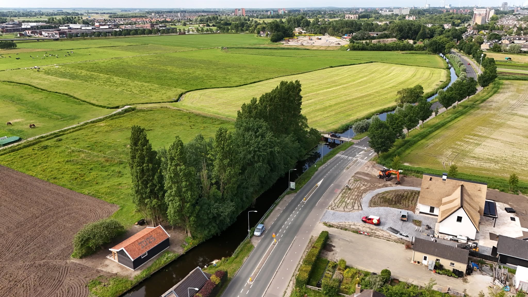 Luchtfoto van groene weilanden met sloot en weg, met huizen en een graafmachine op een bouwterrein