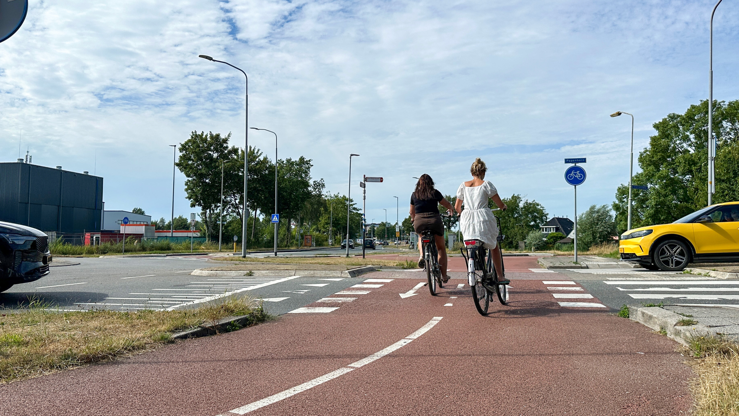 Twee fietsers rijden over een rood fietspad een kruising op, met auto's en verkeersborden op de achtergrond