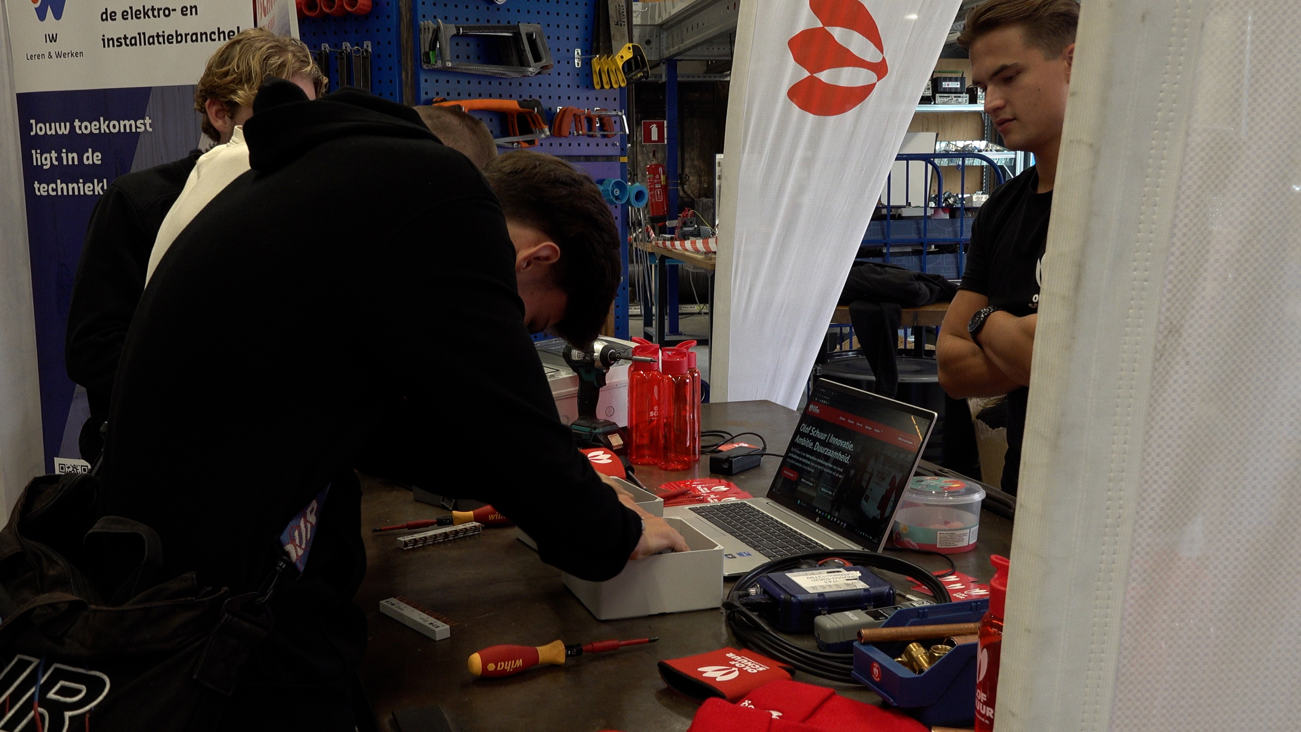 Jongeren bij een techniekstand werken aan een installatie op een werkbank met gereedschap, laptop en rode bidons