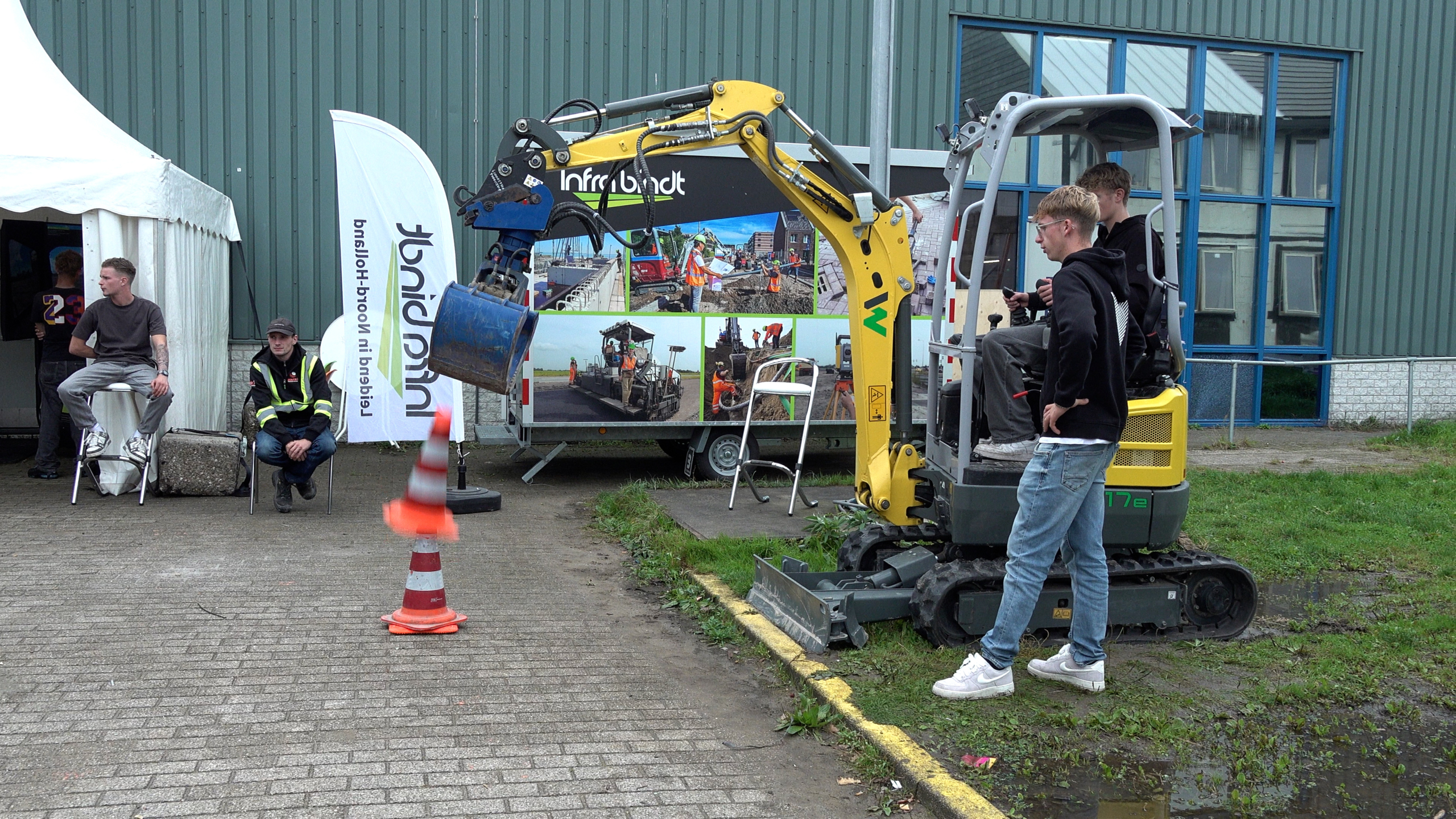 Jongen bedient een minigraafmachine bij een demonstratie, terwijl anderen toekijken naast banners en een tent