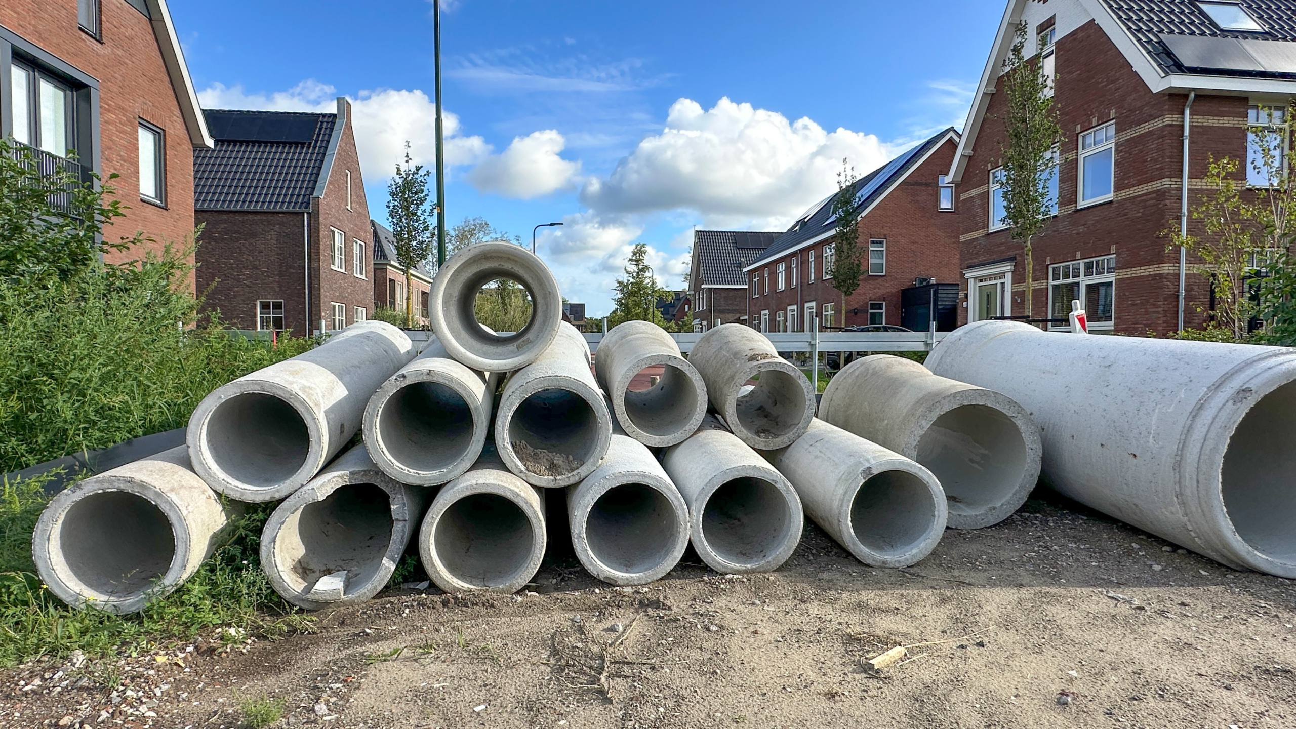 Stapels betonnen buizen liggen langs een straat in een nieuwbouwwijk met bakstenen huizen onder blauwe lucht