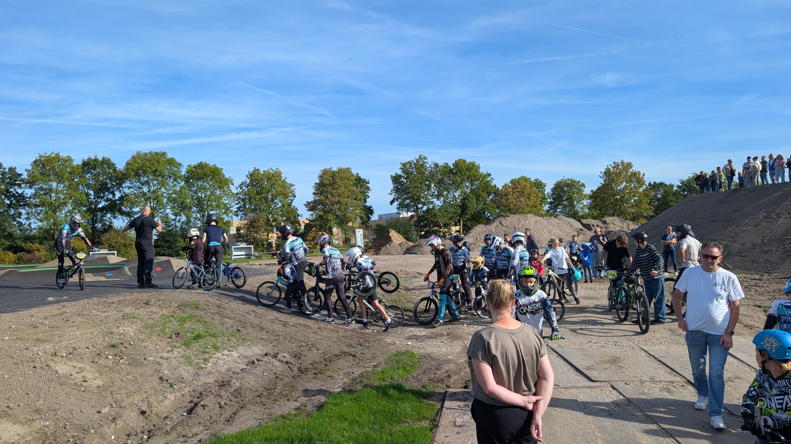 Groep BMX-rijders met helmen en fietsen staat klaar bij een baan, terwijl toeschouwers langs de rand kijken