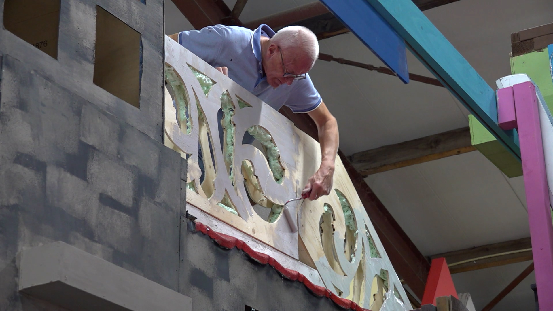 Man met grijs haar schildert een houten decorpaneel met uitgesneden vormen in een werkplaats