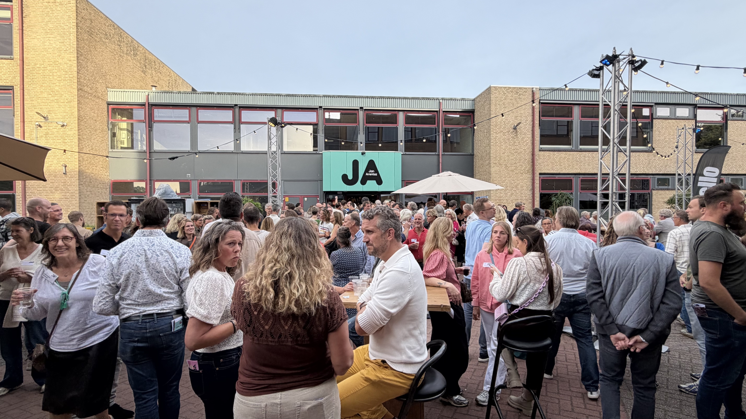 Druk bezocht buitenfeest op binnenplaats, met mensen rond statafels en lichtslingers voor gebouw met JA-bord