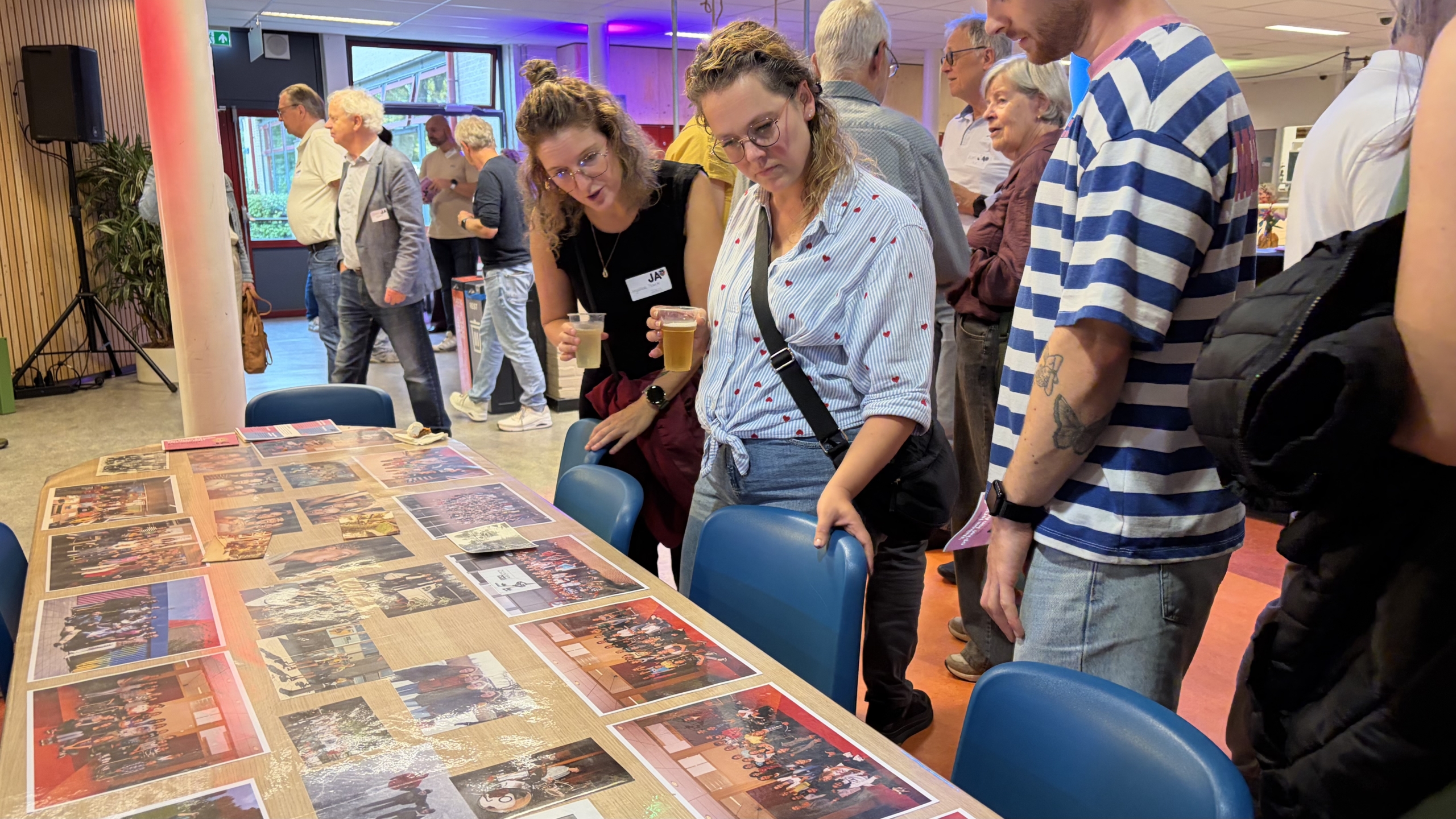 Bezoekers bekijken foto’s op een tafel tijdens een informele bijeenkomst, terwijl twee vrouwen met drankjes meekijken
