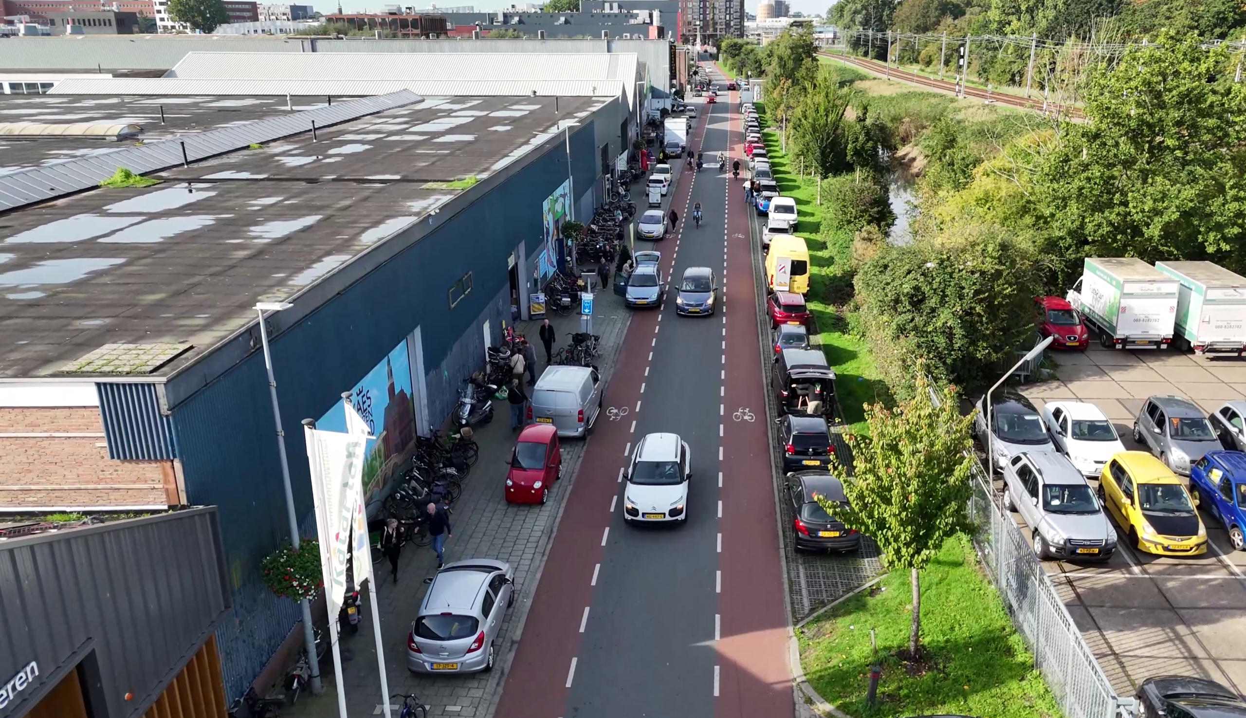 Luchtfoto van een straat langs een groot industriepand, met auto’s, fietsers en bomen langs spoor en water
