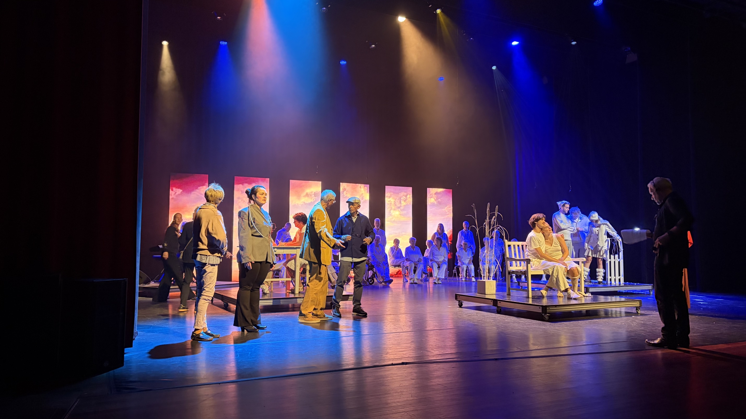 Toneelrepetitie met acteurs onder kleurrijke spotlights, met achteraan een groep in wit en een bankje op het podium