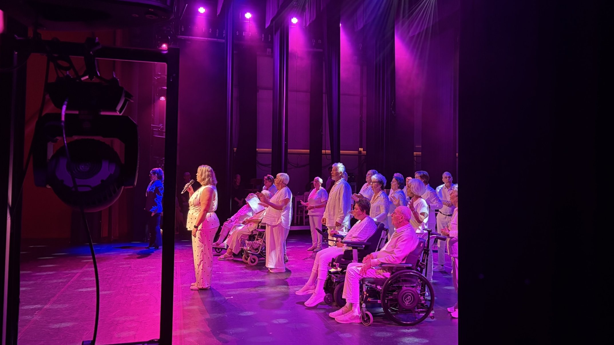 Groep ouderen, sommigen in rolstoel, op toneel onder paarse spotlights terwijl een vrouw met microfoon zingt