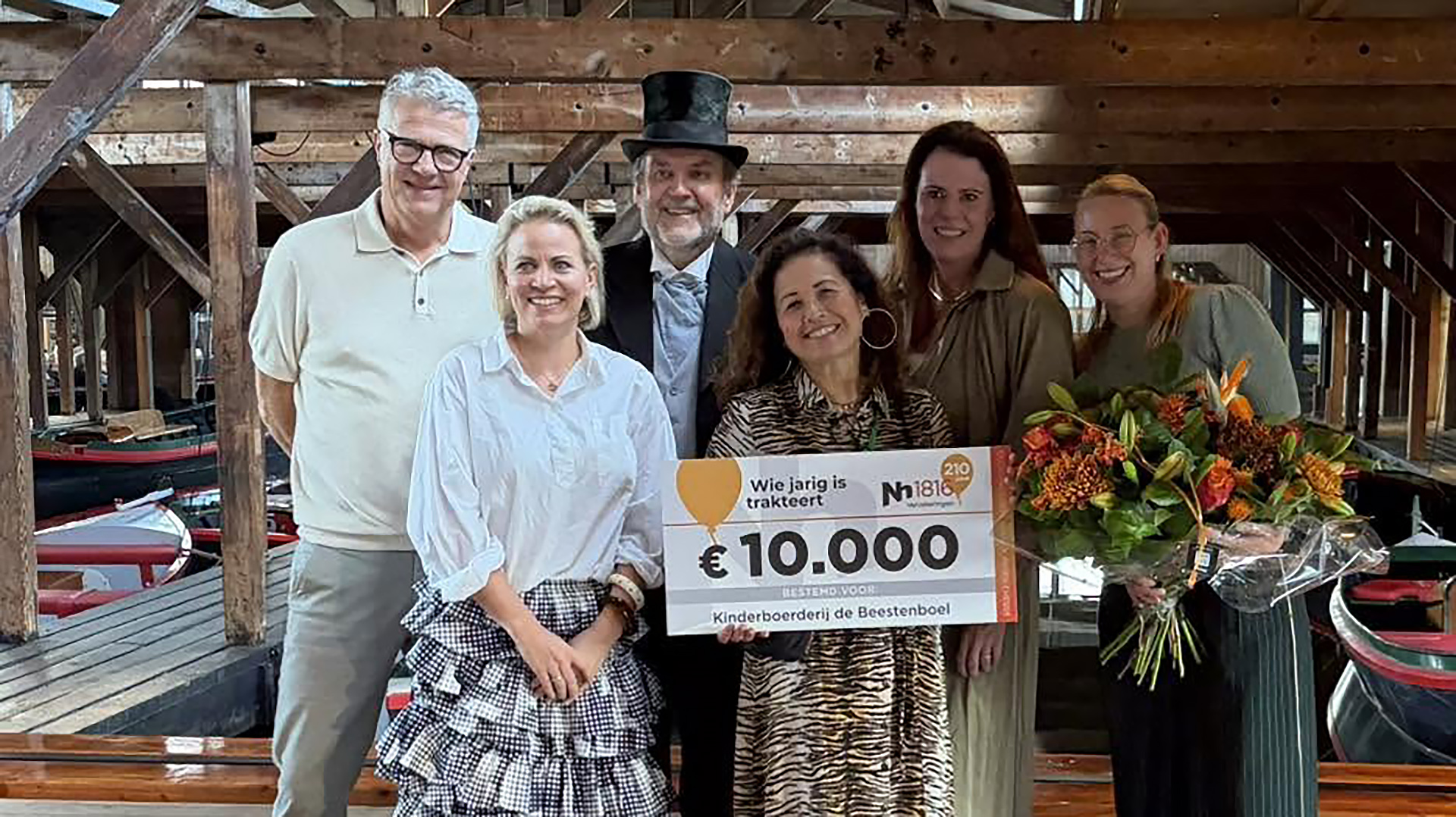 Groep mensen poseert in houten loods met cheque van €10.000 en een bos bloemen