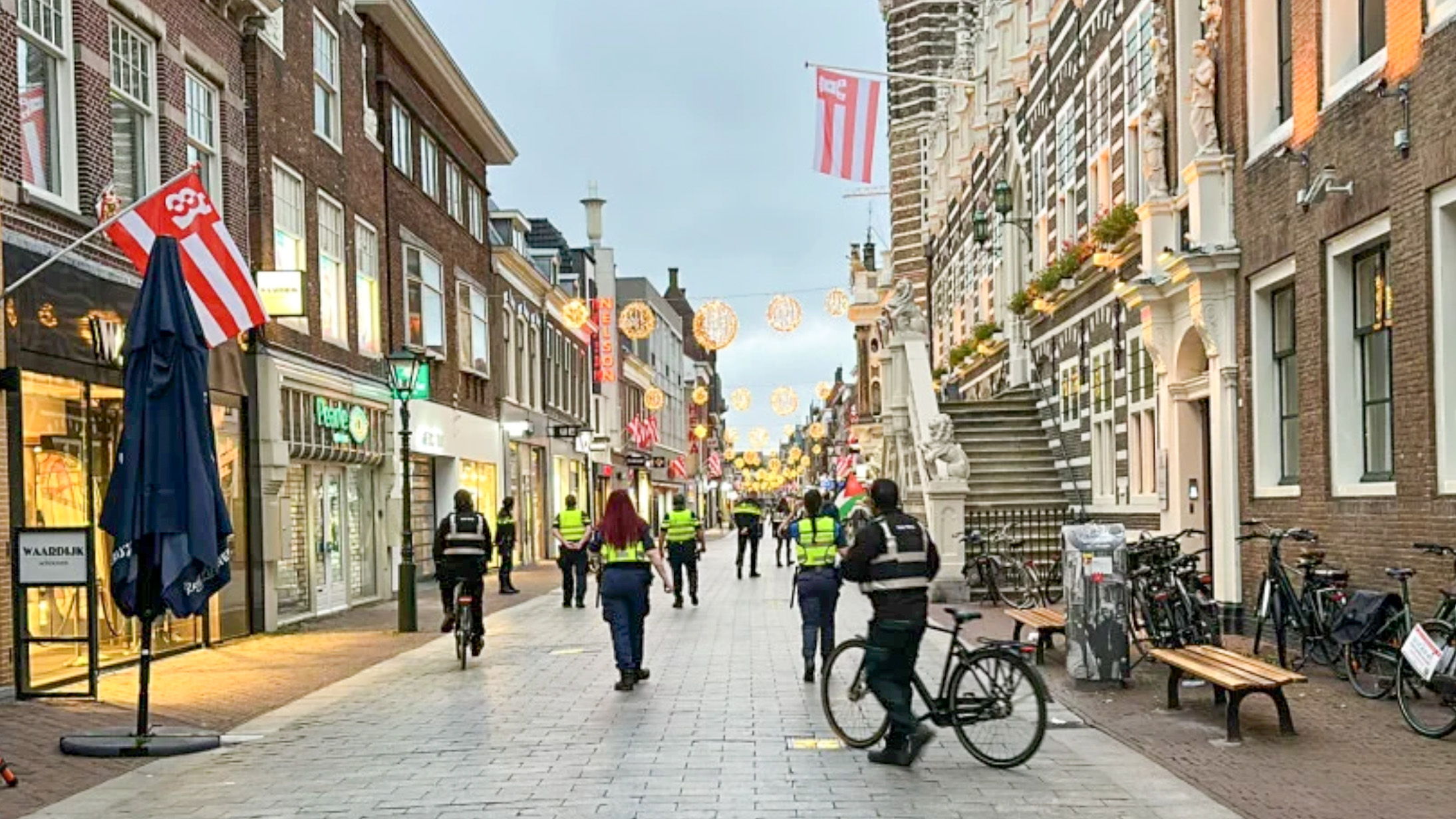 Winkelstraat met feestverlichting; agenten en handhavers lopen en fietsen langs winkels en gevels met vlaggen