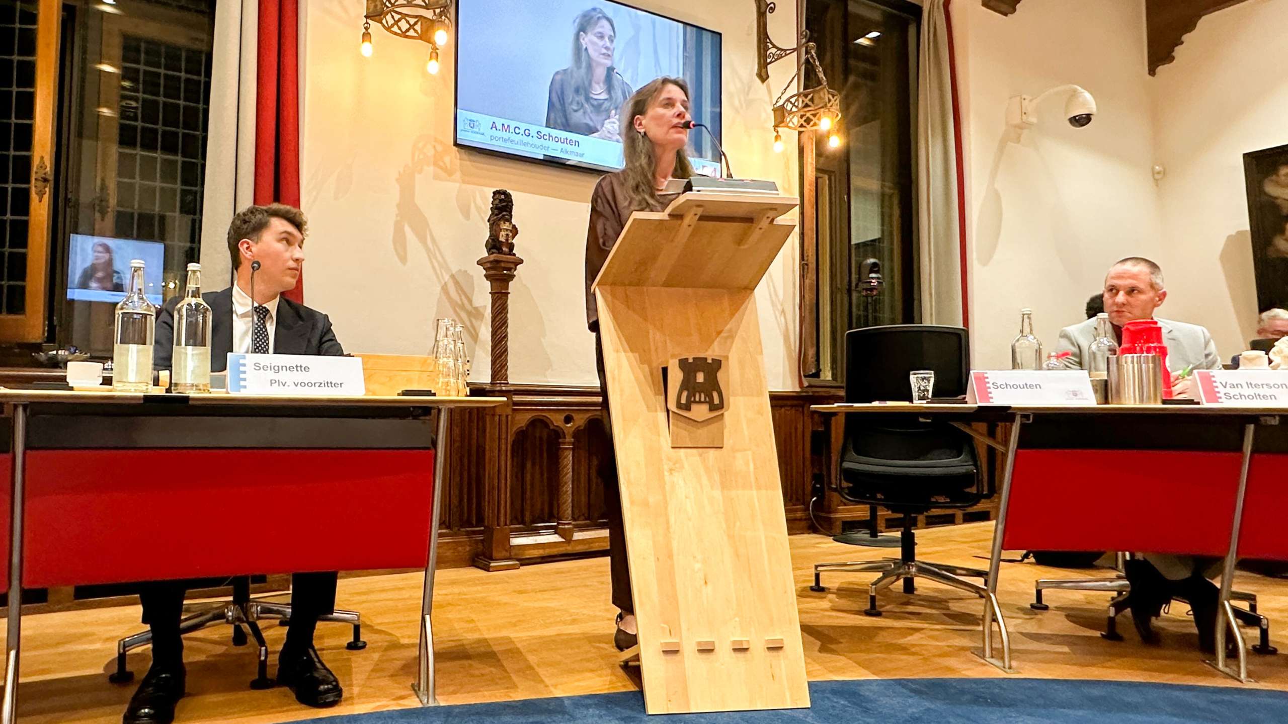 Vrouw spreekt vanaf een spreekgestoelte tijdens raadsvergadering, met scherm erboven en raadsleden aan tafels met naamkaartjes