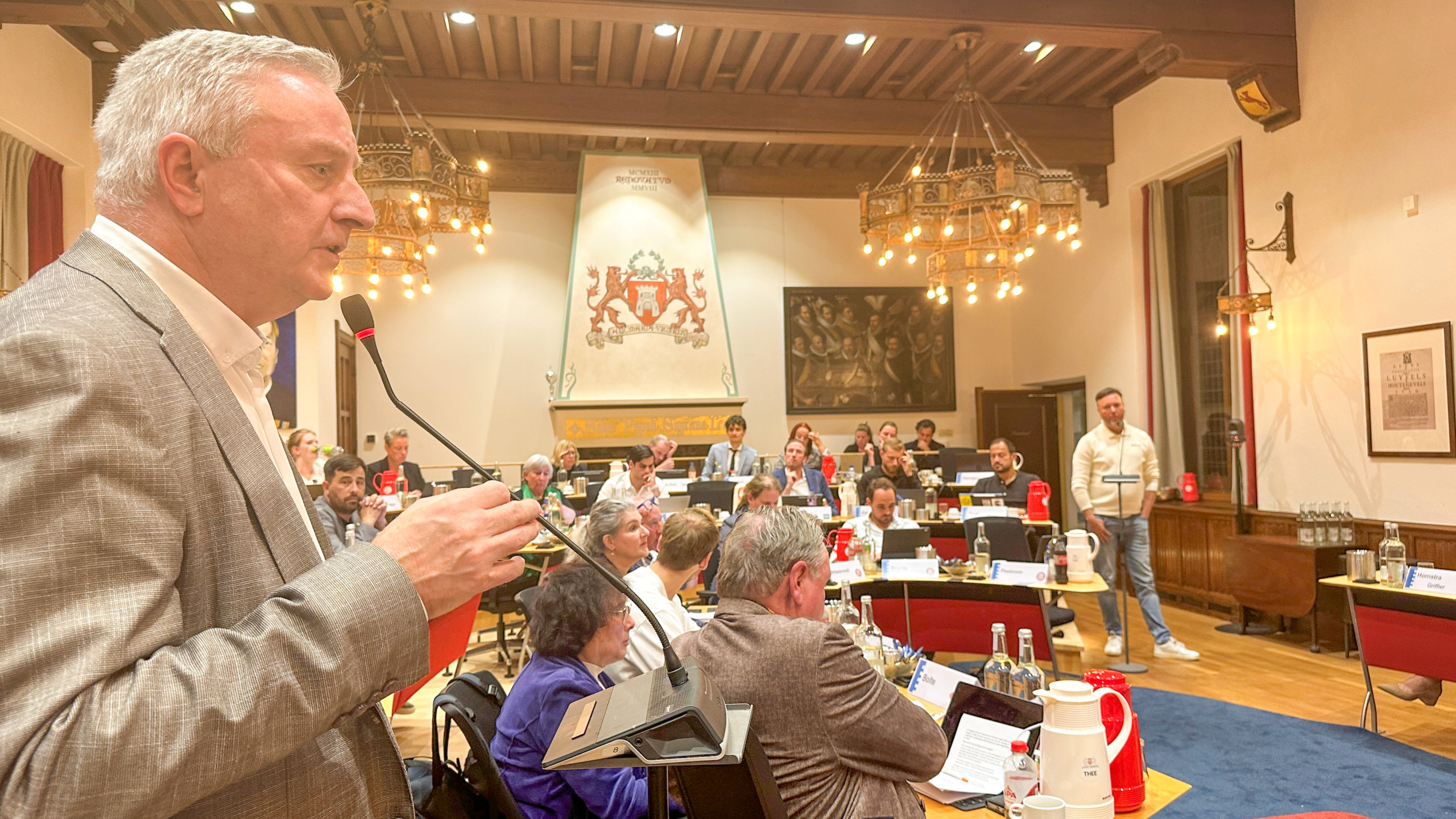 Man spreekt in een raadszaal in een microfoon, terwijl aanwezigen aan tafels met laptops en documenten luisteren
