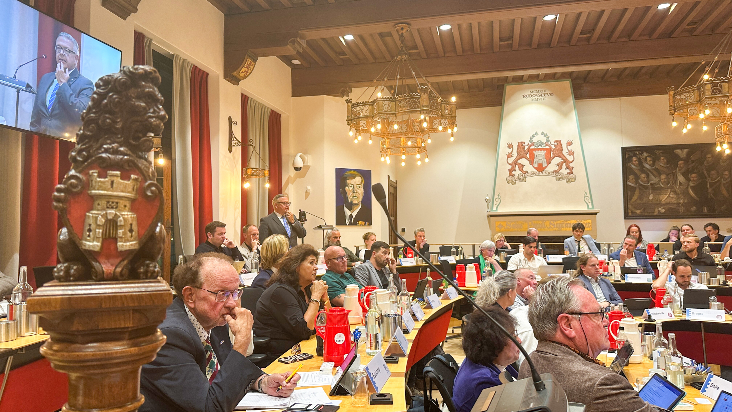 Raadsvergadering in historische zaal; spreker aan lessenaar, publiek aan tafels, stadswapen en groot scherm op de achtergrond