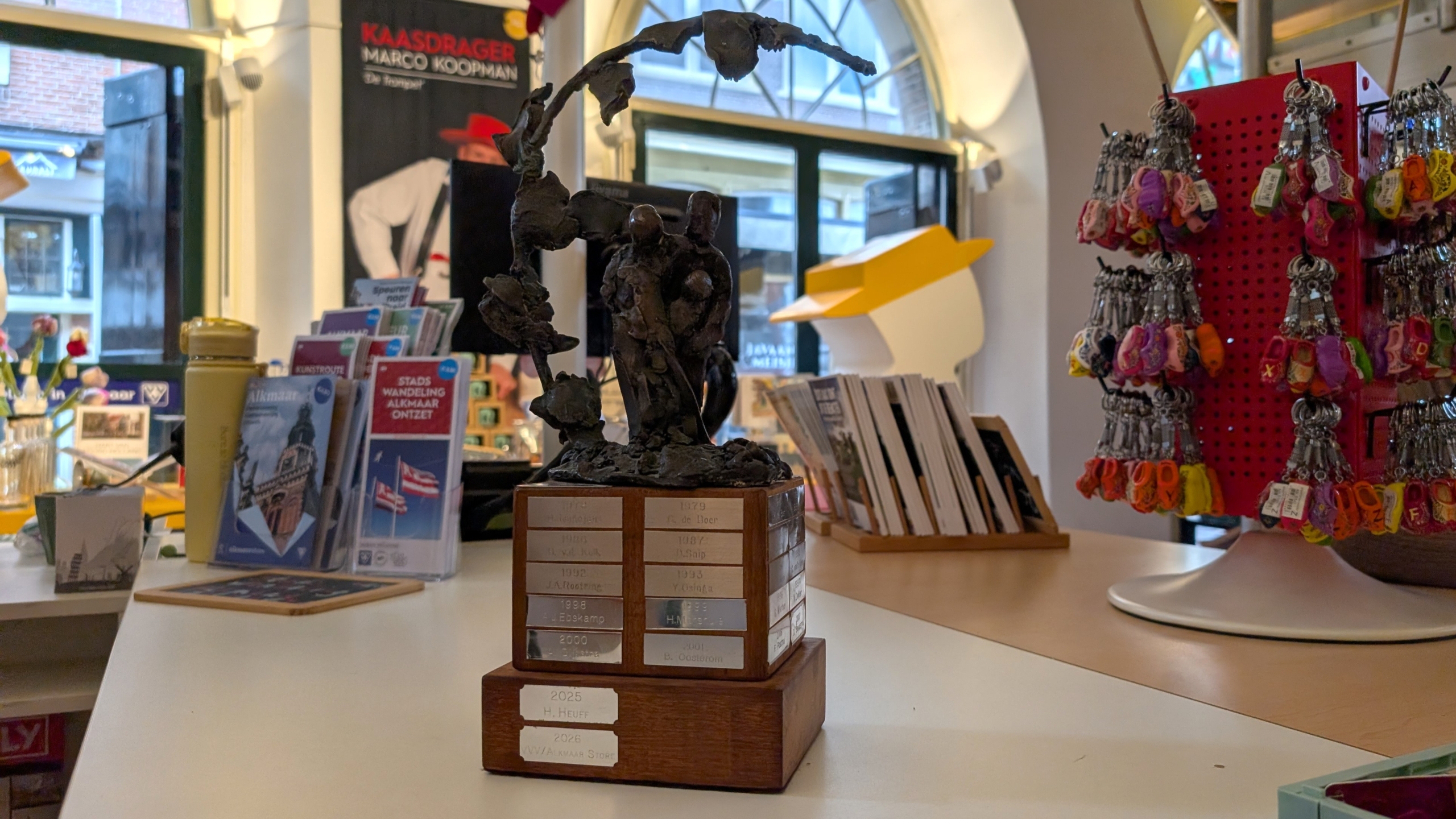 Bronzen beeldje op houten trofeevoet op balie, met reisbrochures en kleurrijke sleutelhangers in souvenirwinkel
