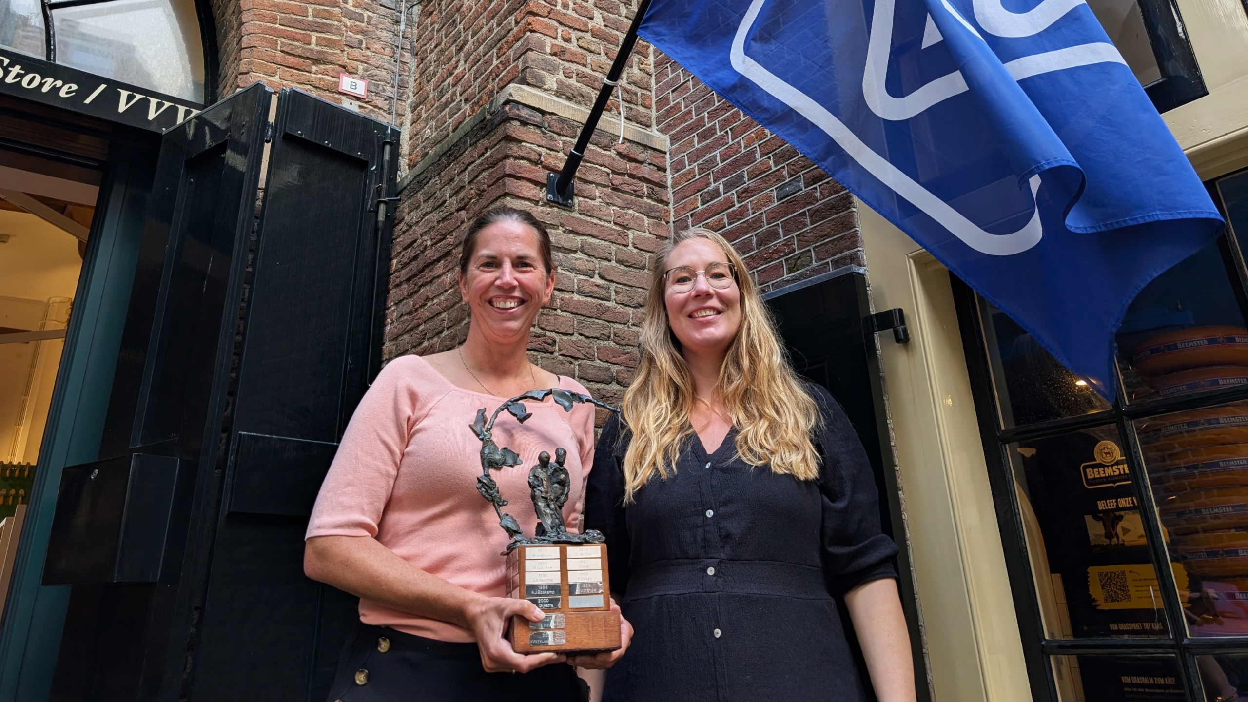 Twee lachende vrouwen staan voor een bakstenen gevel en houden samen een trofee vast, met een blauwe vlag rechts