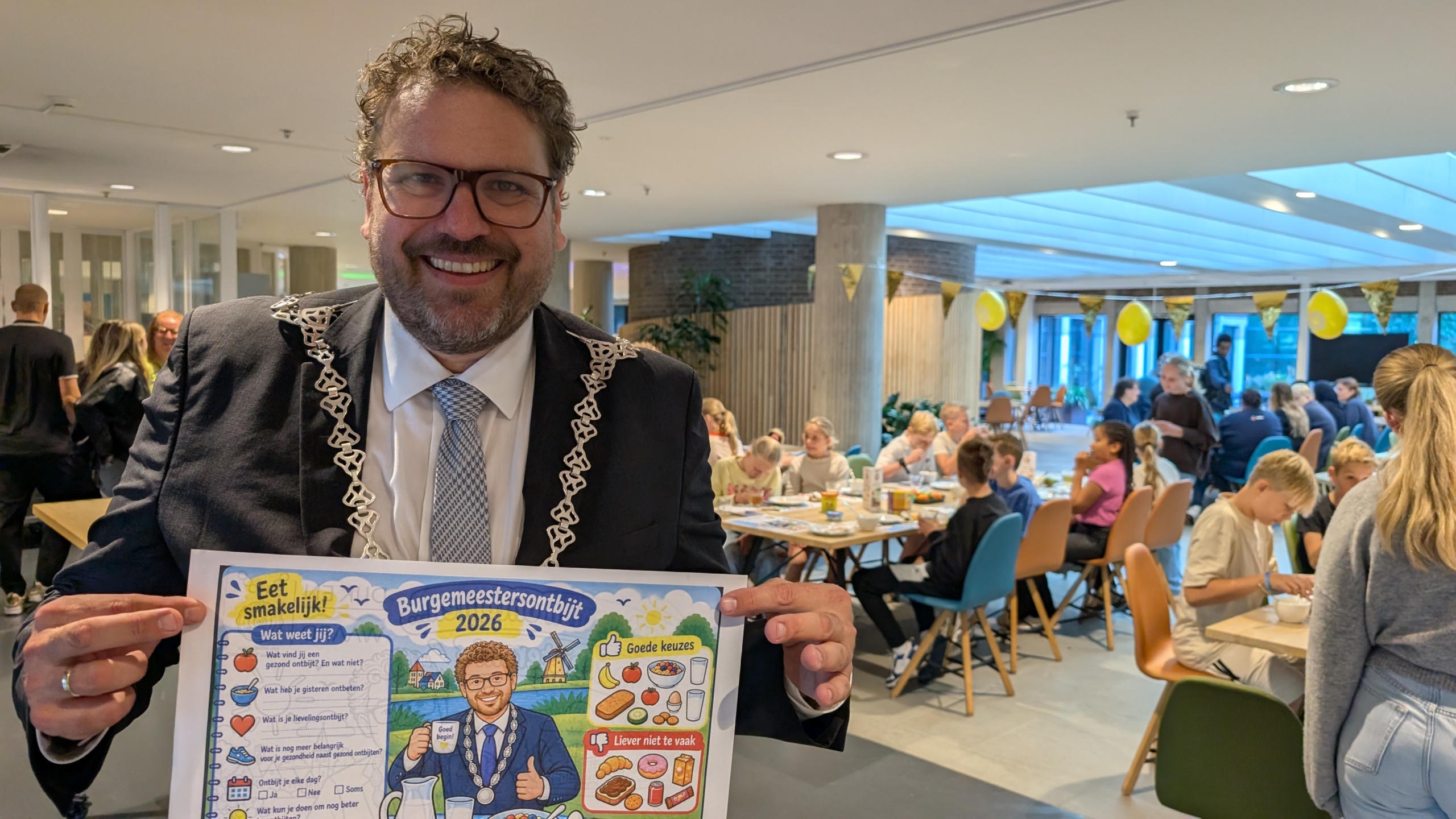 Man in pak met ambtsketen houdt poster “Burgemeestersontbijt 2026” vast in een drukke zaal met ontbijttafels