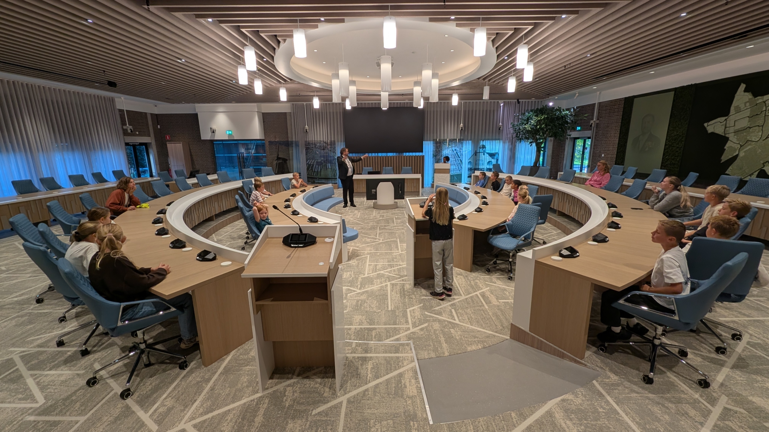 Kinderen en begeleiders in een moderne raadszaal met halfronde tafels, terwijl een spreker in het midden uitlegt