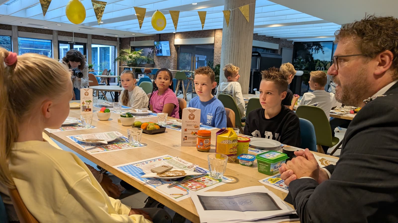 Kinderen zitten aan een lange tafel met bordjes en eten, terwijl een man in pak met hen in gesprek is in een kantine