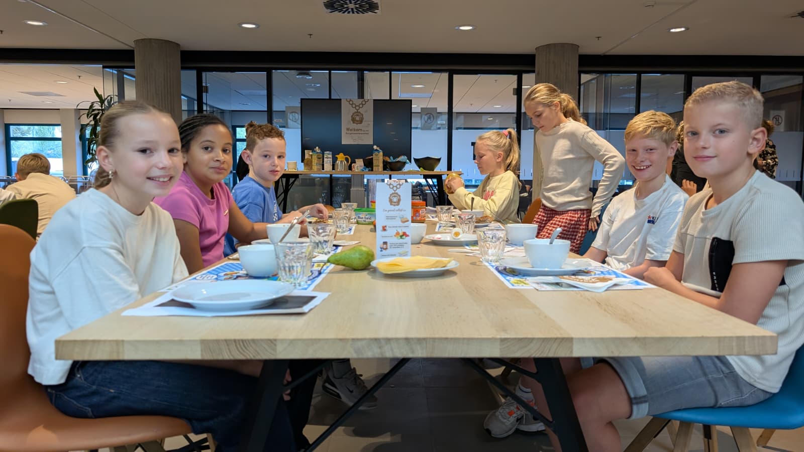 Groep kinderen zit aan een lange tafel met ontbijtspullen, borden en glazen in een eetruimte