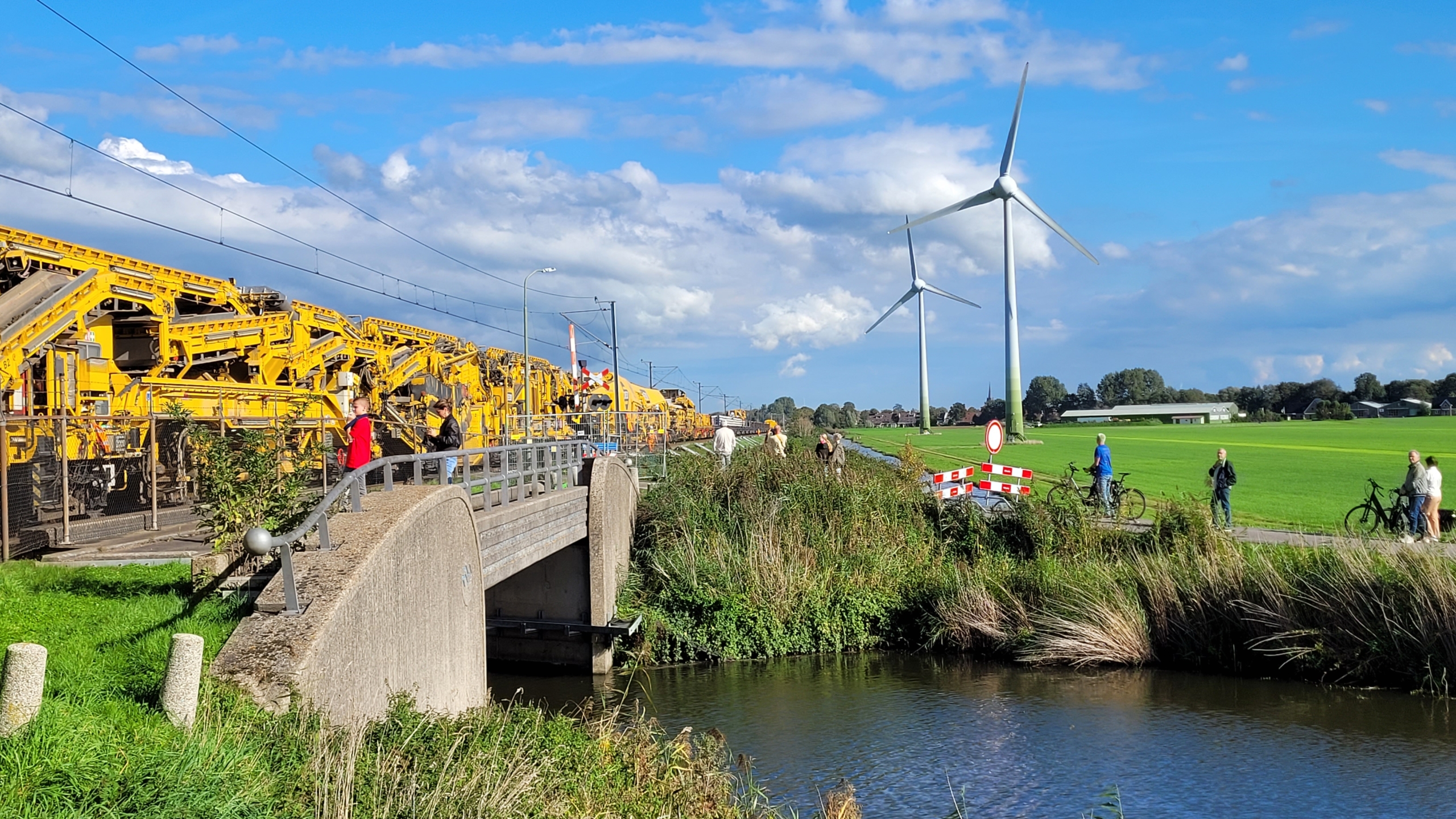 Gele spoorwerkmachines langs het spoor naast een brug over een sloot, met toeschouwers en windturbines in de polder