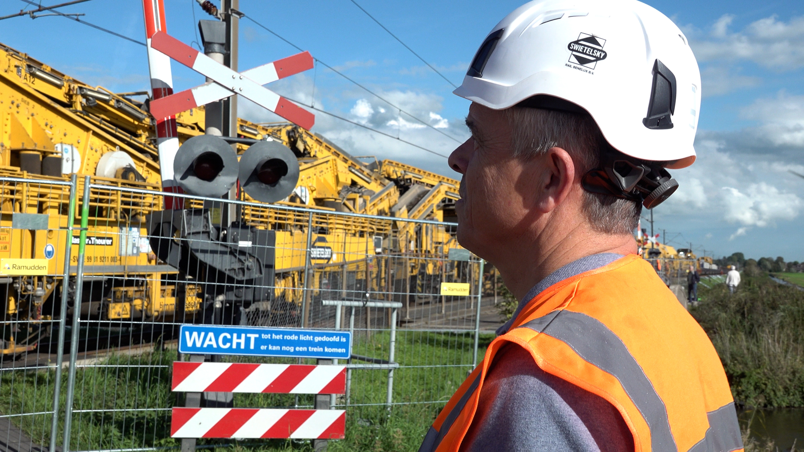 Spoorwerker met witte helm en oranje veiligheidshesje bij spoorwegovergang met bord WACHT en gele werkmachines